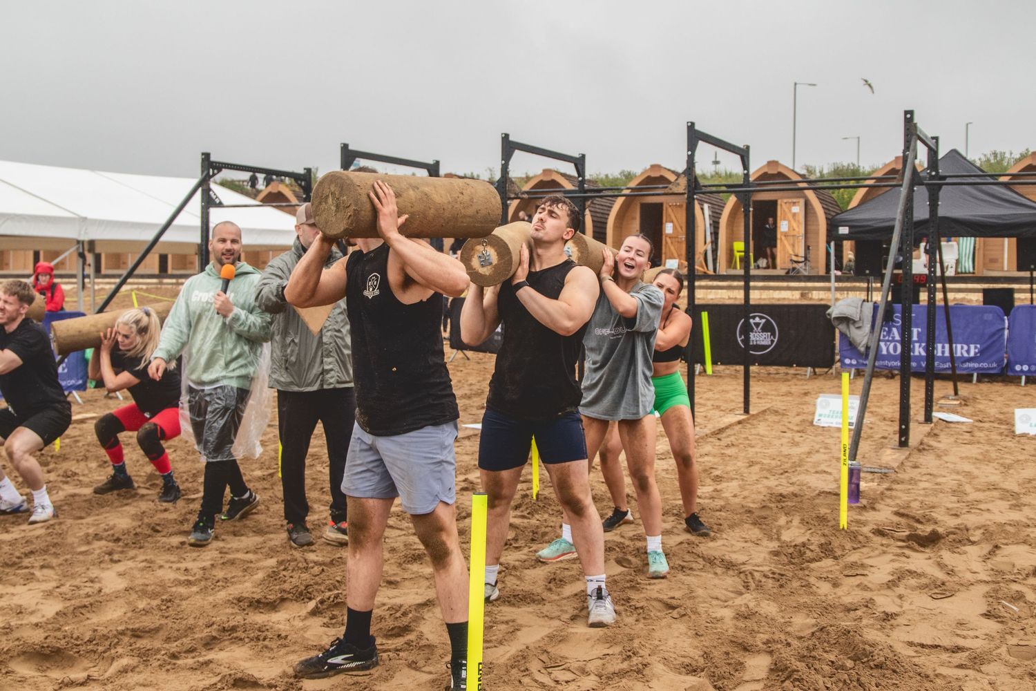 Athletes compete in an intense log lifting competition at an outdoor sand arena during a strength sports event.
