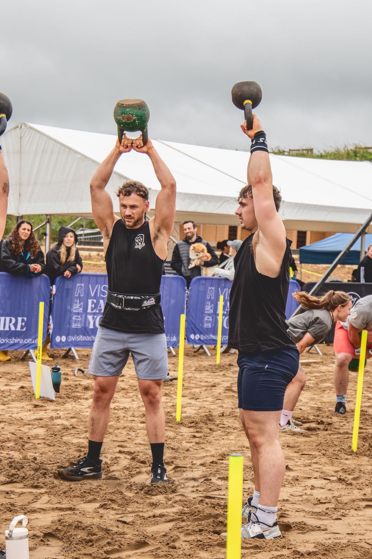 Two athletes perform synchronized kettlebell lifts during an outdoor competition on sandy ground with event banners visible.