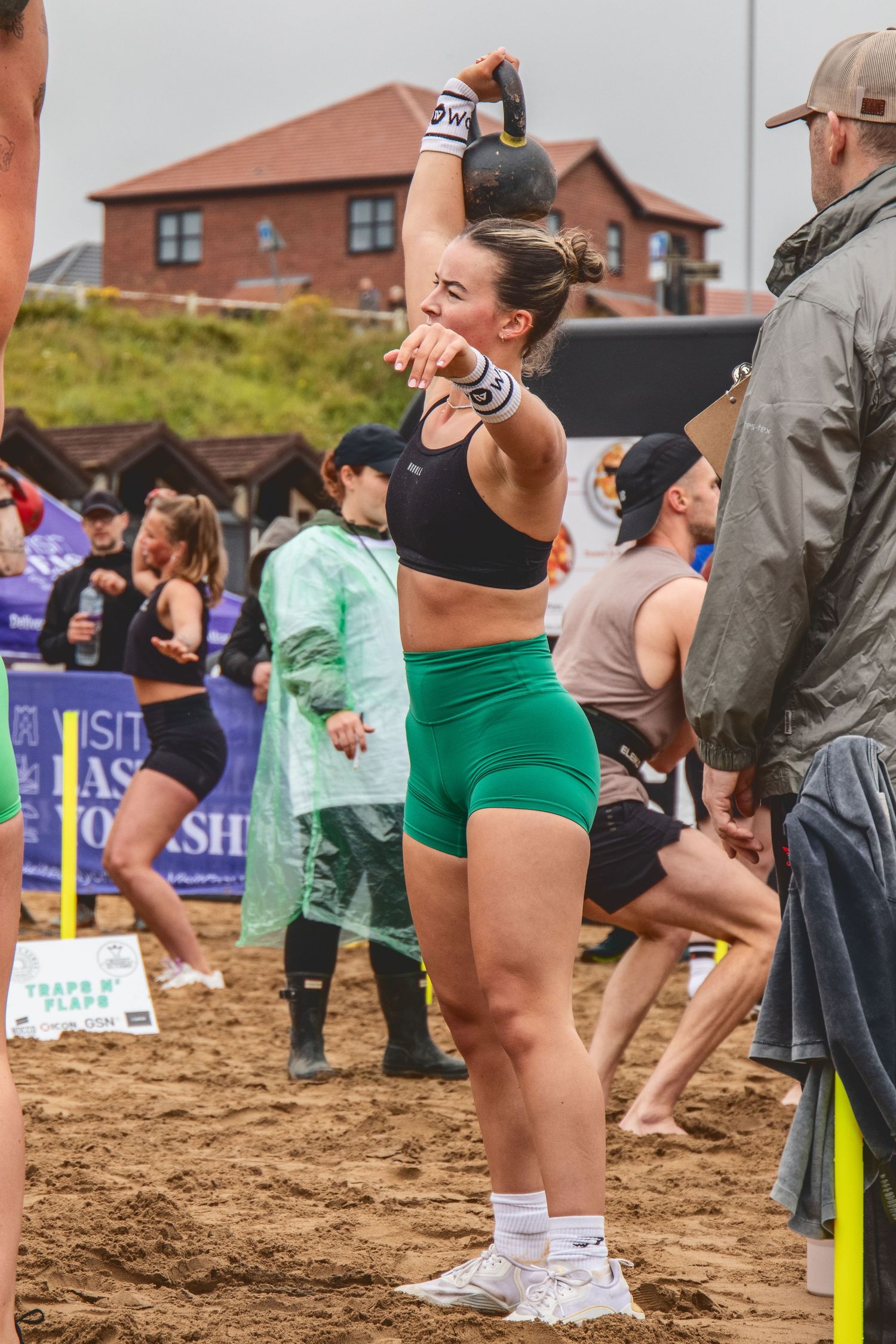 An athlete in a black sports bra and green shorts stretches outdoors during a competitive event on sandy ground.