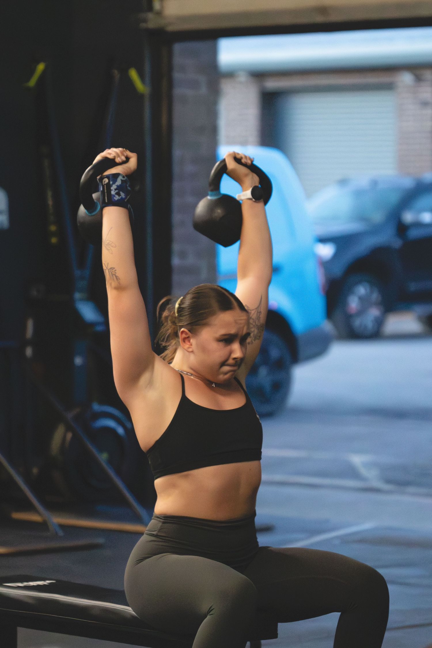Athletic person in black sports bra and leggings performing overhead kettlebell exercise at a gym.
