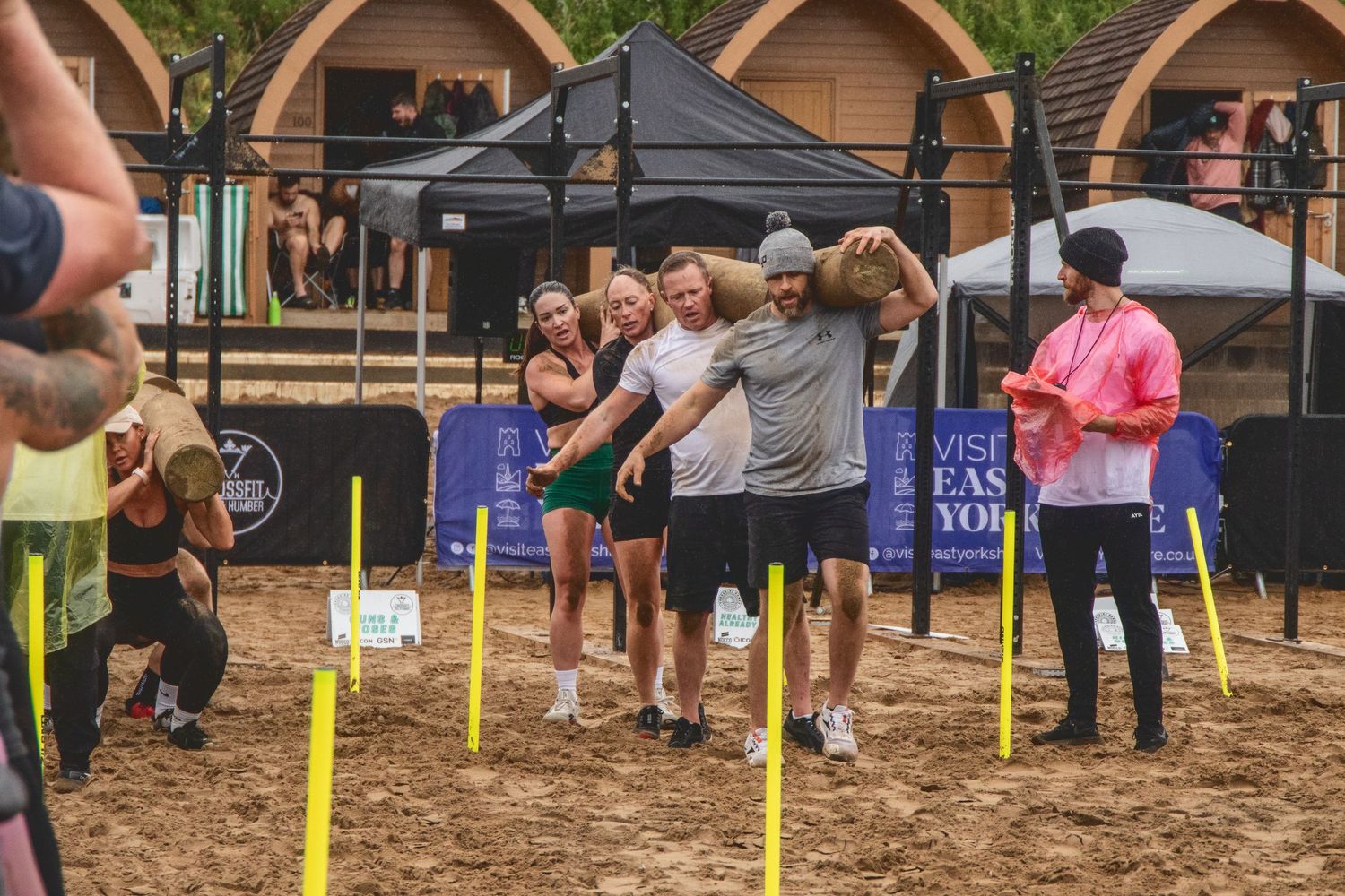 A group of people participating in an outdoor game event with yellow position markers on sandy ground under pop-up canopies.