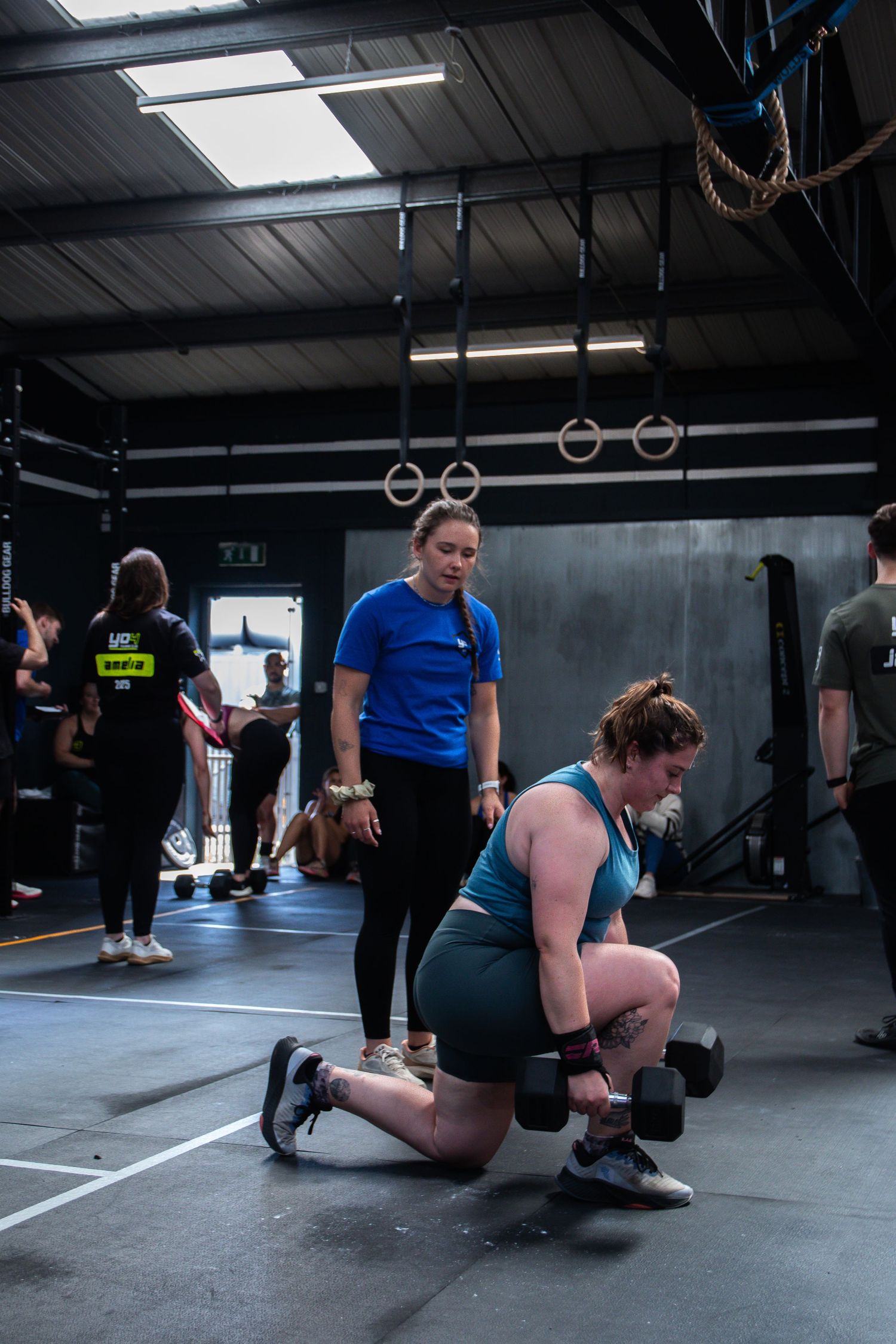 Athletes training in a CrossFit gym with gymnastics rings and weightlifting equipment visible in the background.