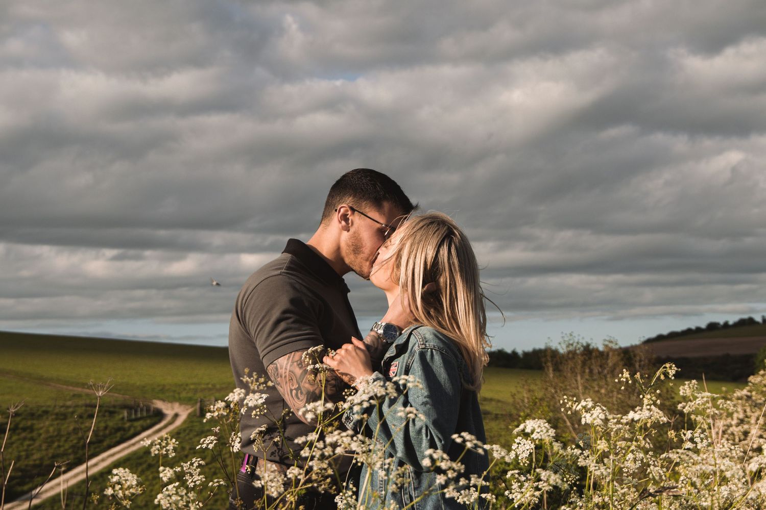 Loving couple sharing kiss in flowering field with dramatic stormy clouds above.