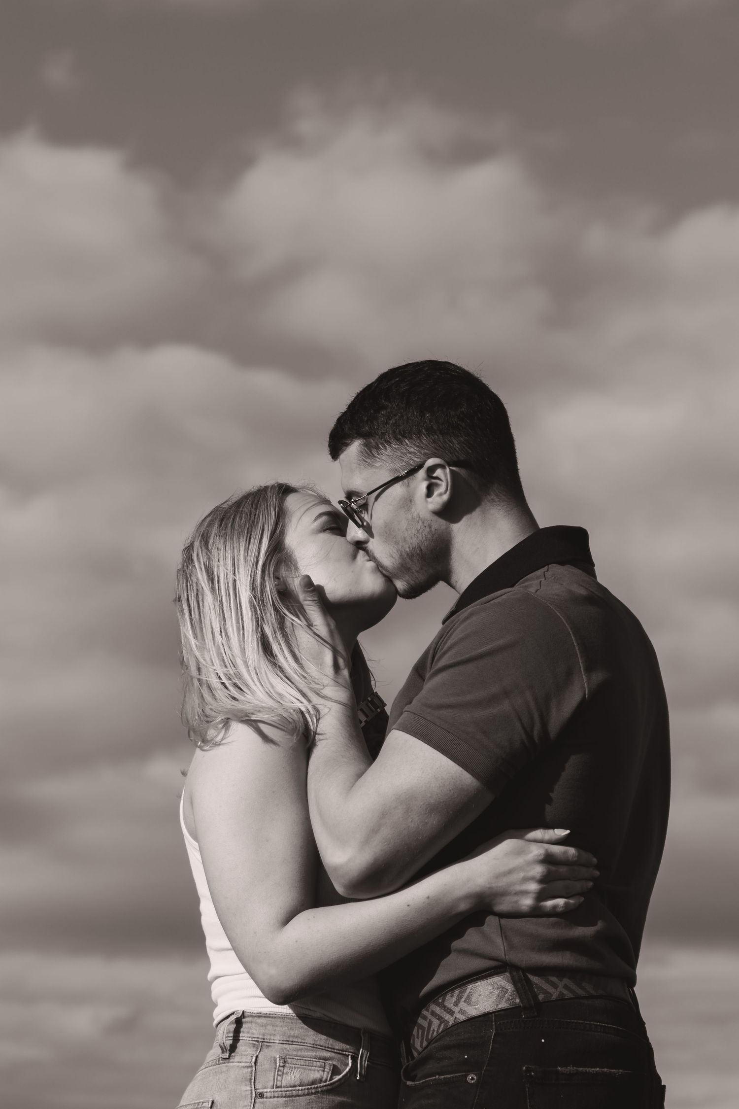 Black and white photo of a couple sharing an intimate kiss against dramatic clouds.