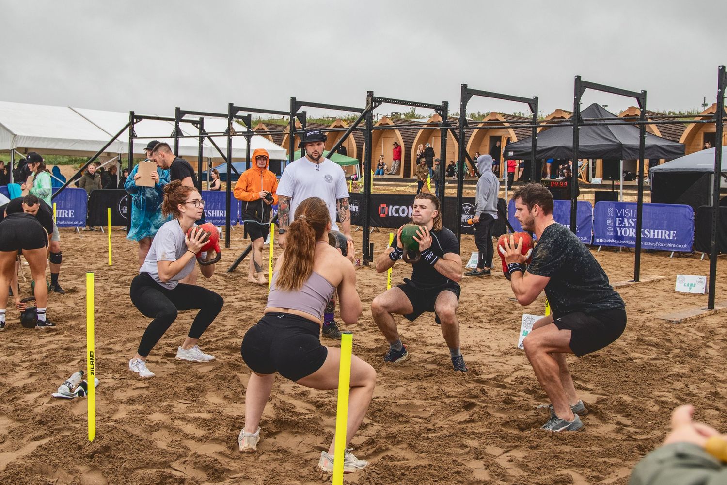 Athletes perform agility drills on sand with yellow training poles during an outdoor fitness competition.