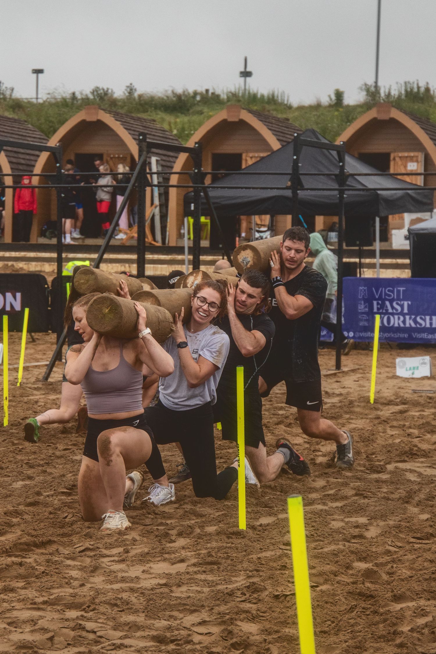 A group works together to carry sandbags during an outdoor team challenge event on a muddy course with arched structures.