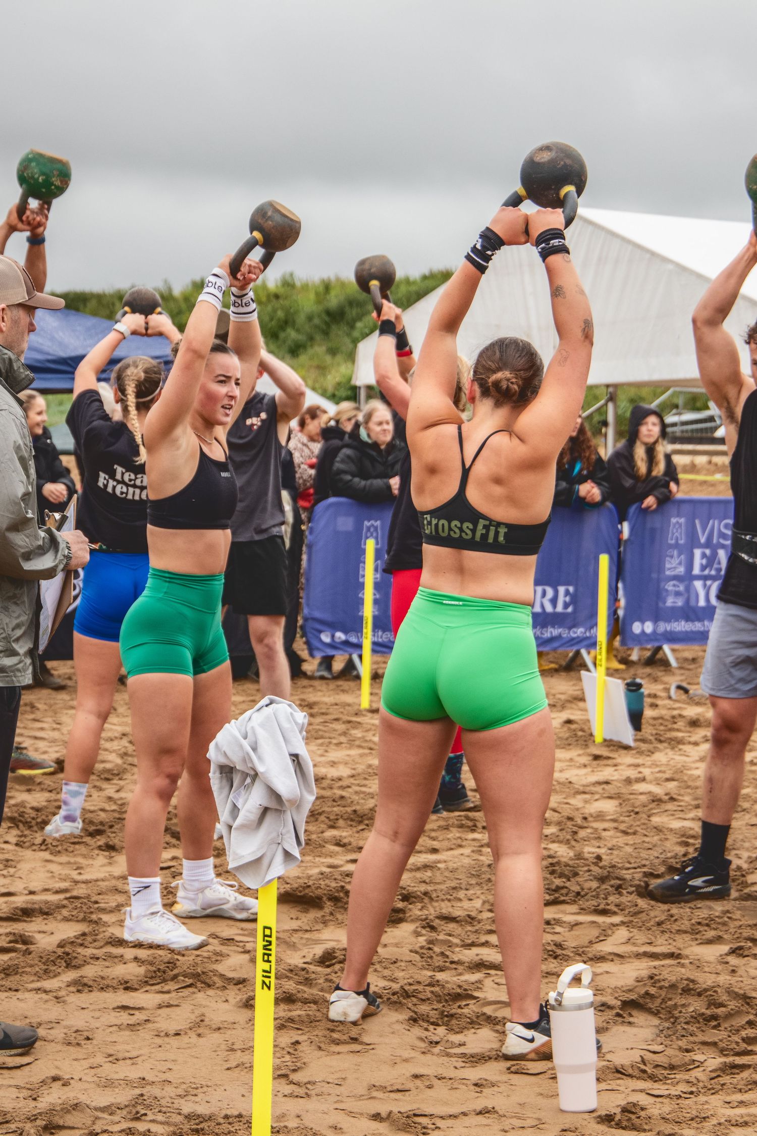 Athletes in green shorts perform synchronized overhead medicine ball movements on a sandy beach during a fitness event.