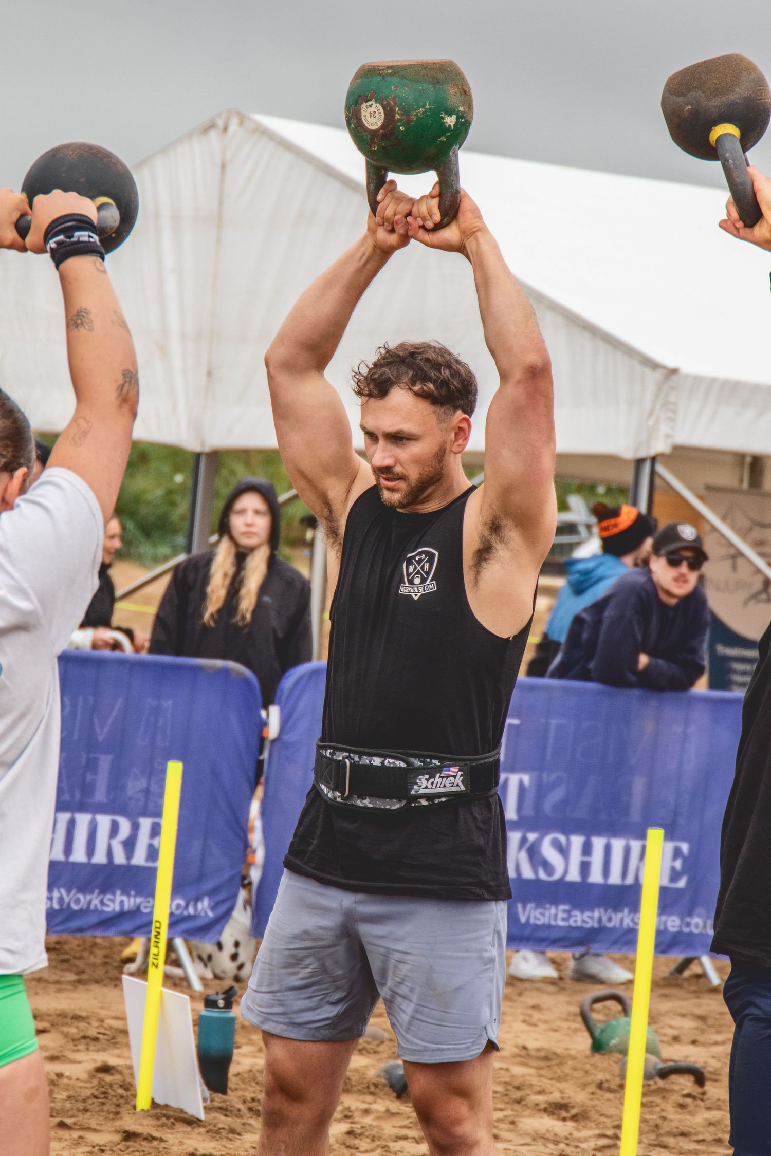 Athletes lift heavy kettlebells during an outdoor strongman competition on a sandy beach with event banners visible.