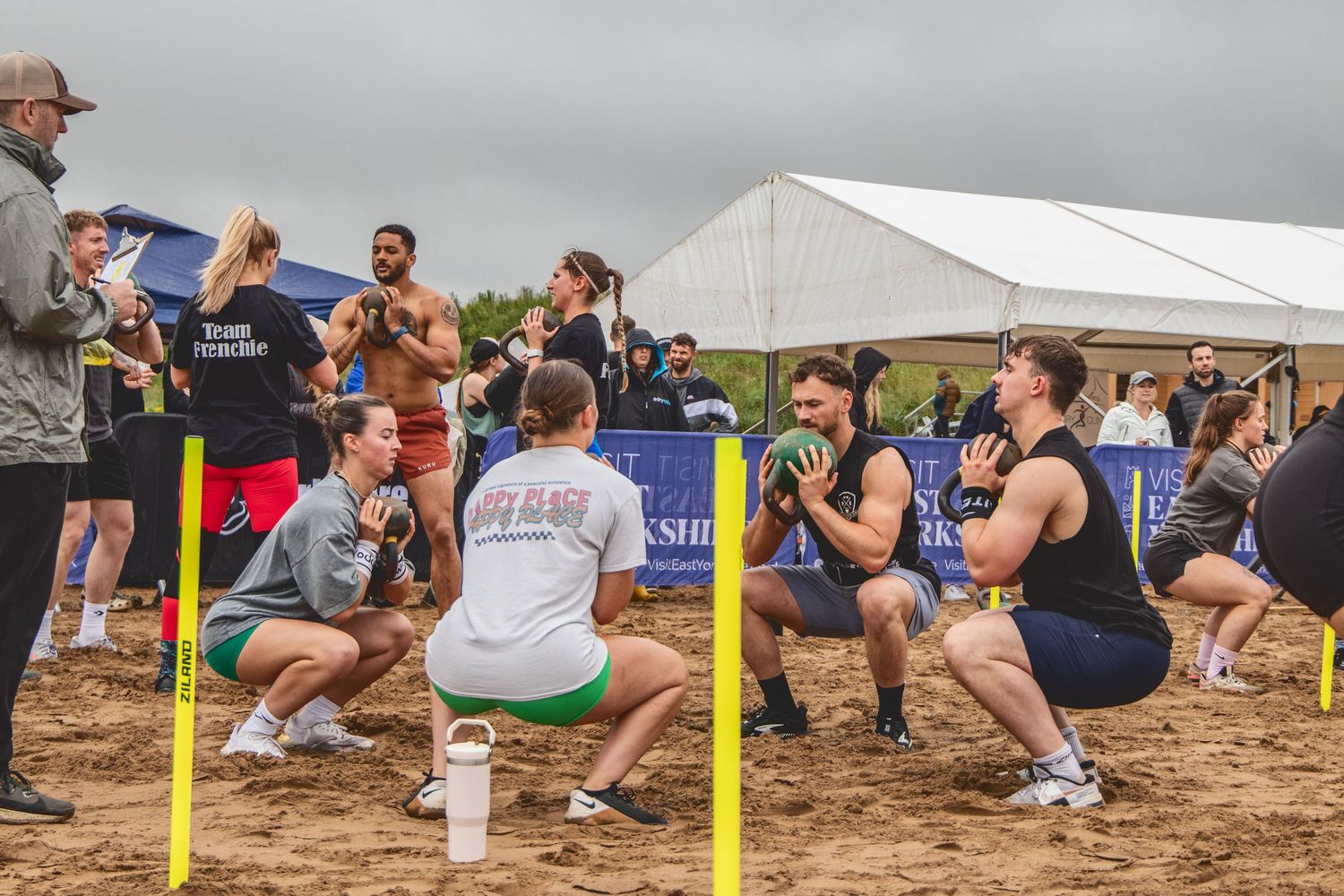 Athletes perform synchronized squats during an outdoor fitness competition on sandy terrain next to a white tent.