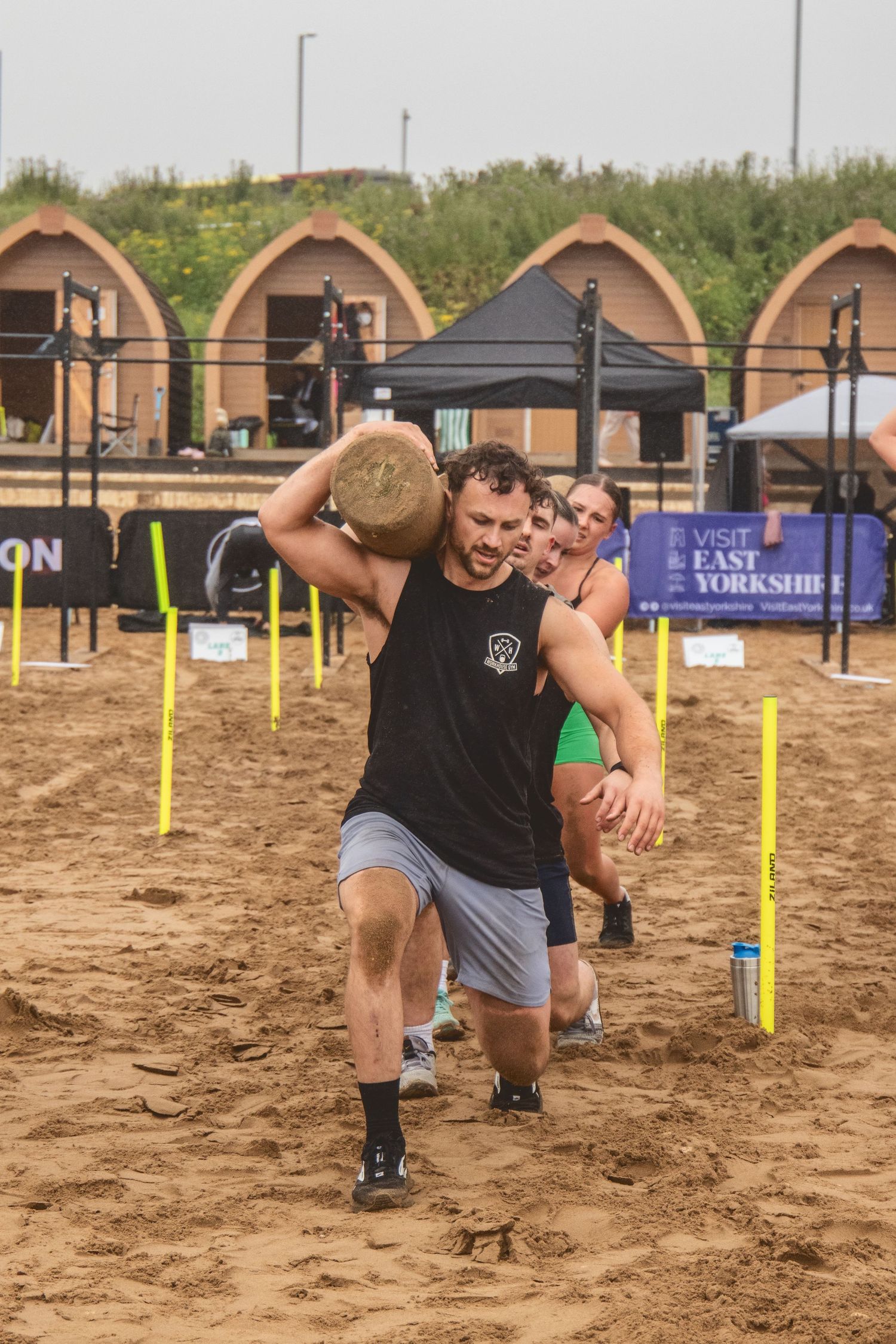 Athletes compete in a strength and conditioning challenge carrying sandbags through yellow markers on sandy terrain.