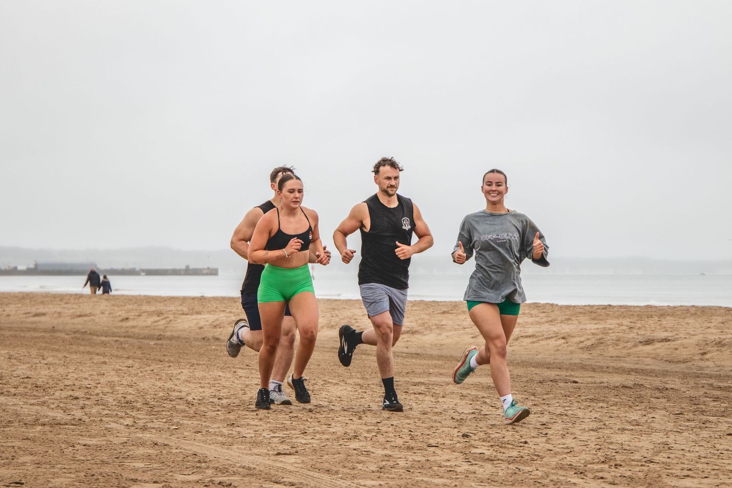 Three people jogging together on a sandy beach during an overcast day with a dog visible in the background.
