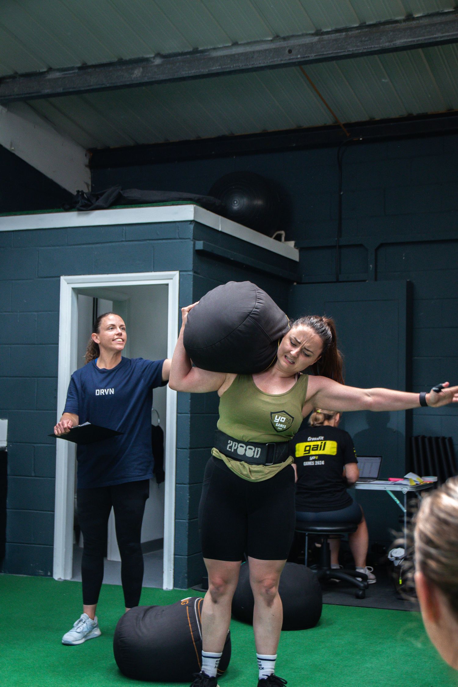Fitness instructor demonstrates proper form for kettlebell swing exercise on green turf flooring in gym studio.