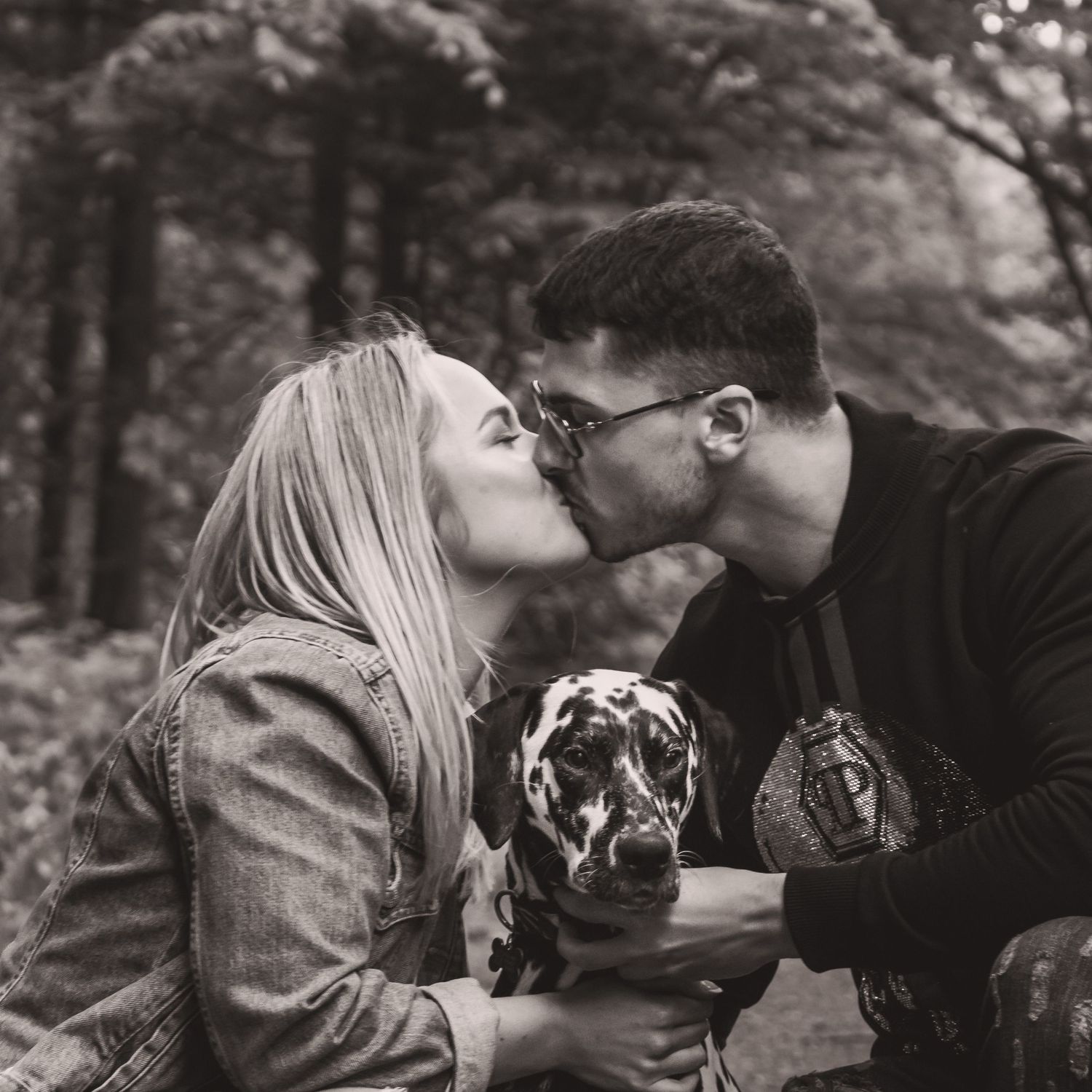 Sweet moment between a couple sharing a kiss while holding their spotted dog in the woods.