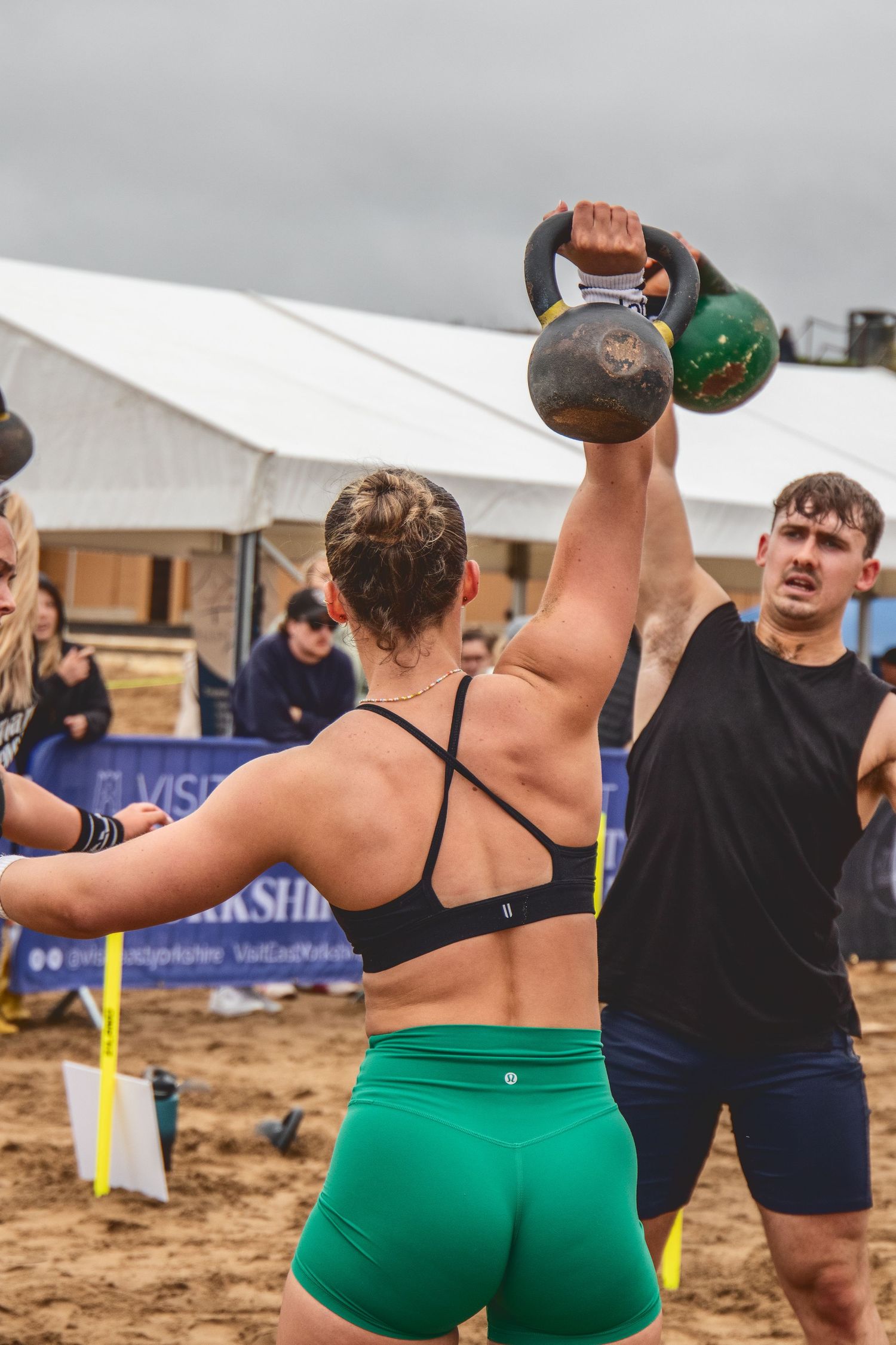 Athletes perform kettlebell exercises during an outdoor fitness competition on a sandy beach.