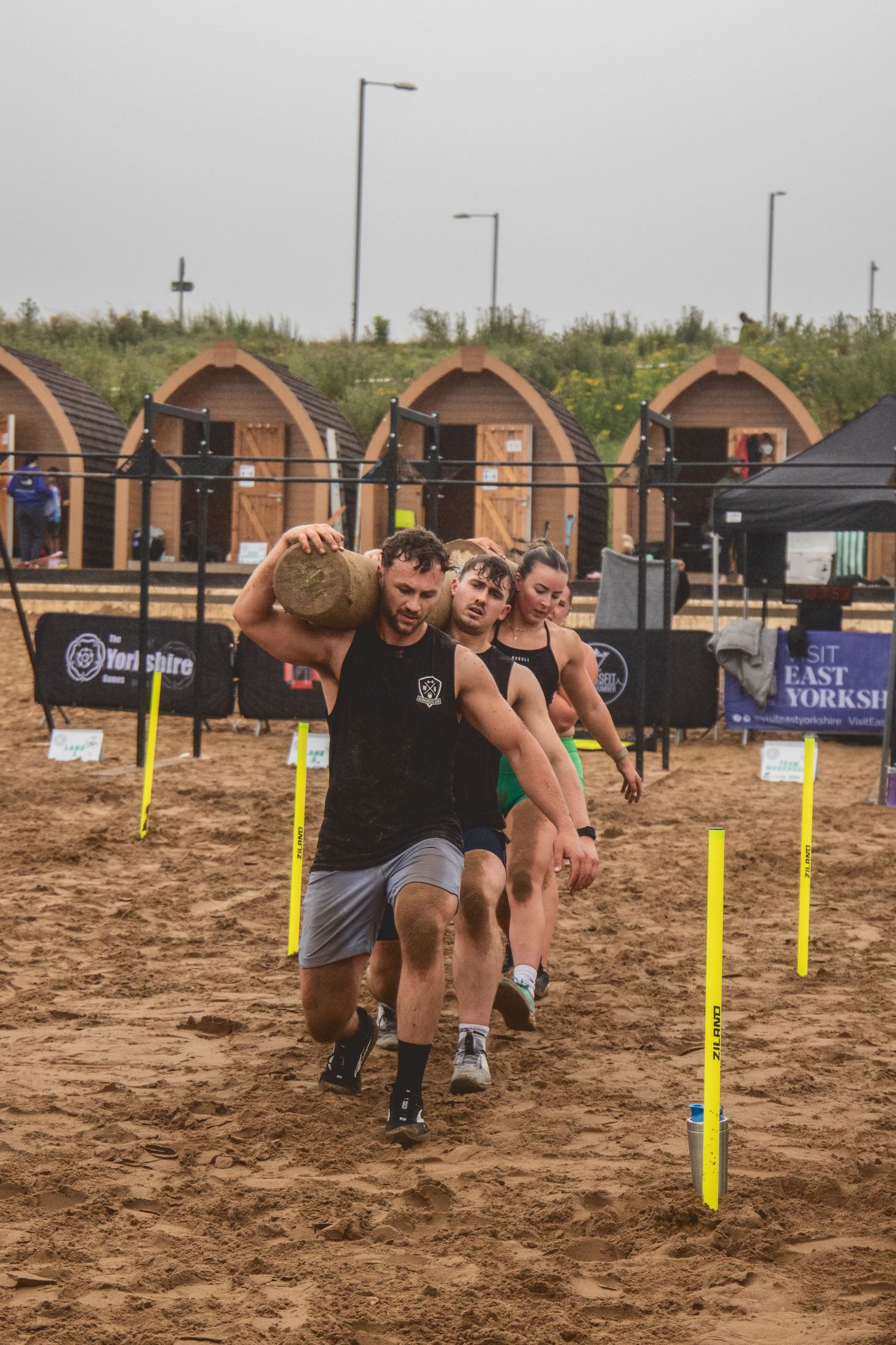 Athletes compete in an outdoor obstacle course on sandy terrain featuring yellow markers and wooden obstacles.