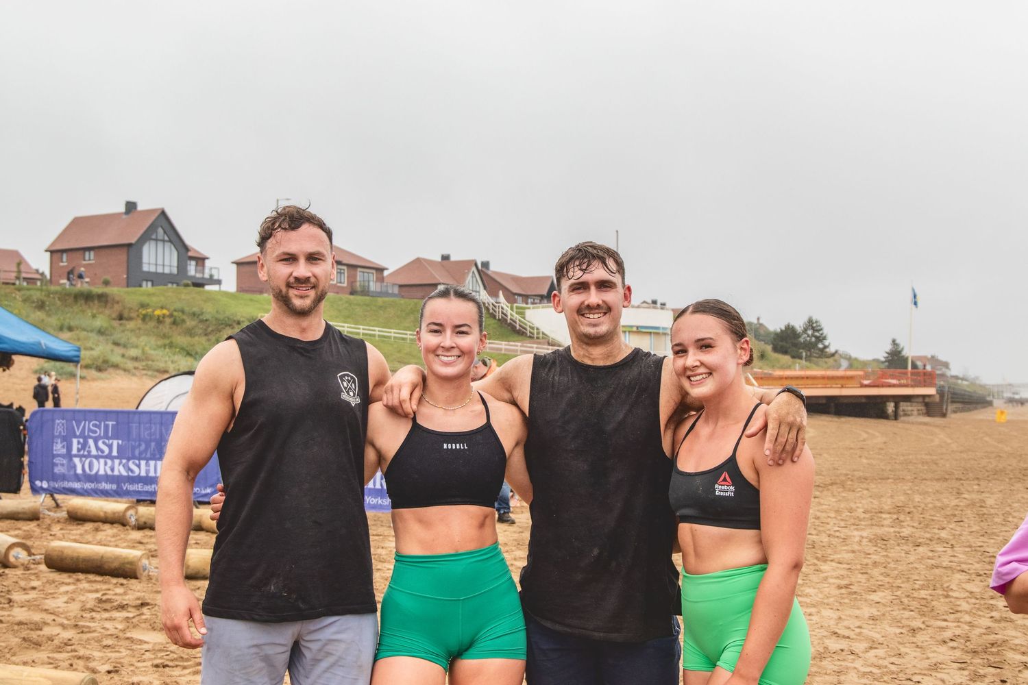 Four athletic friends in workout attire smile together on a beach with houses visible on the hillside behind them.