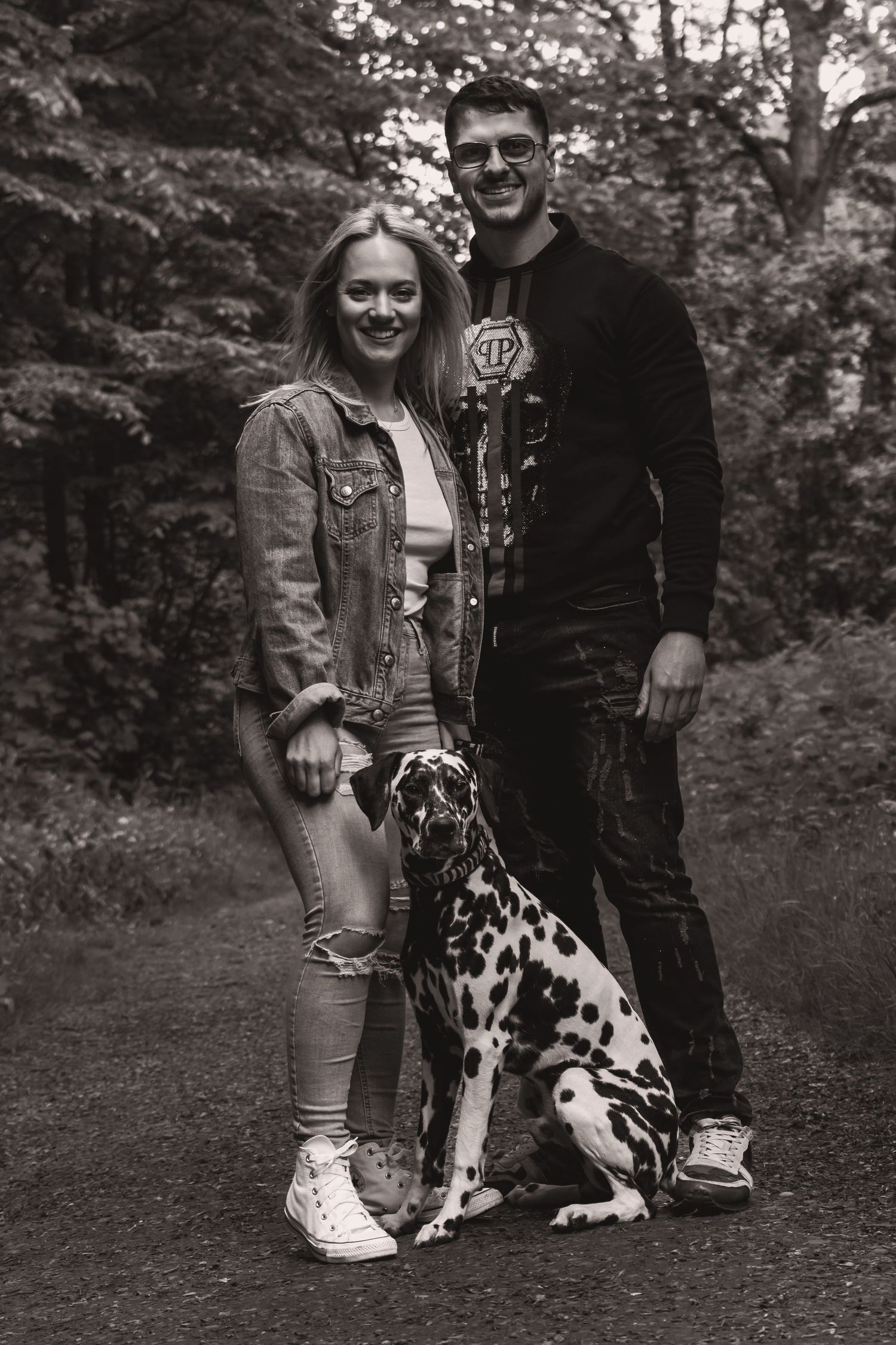 A couple and their Dalmatian dog pose together on a wooded path in black and white photography.