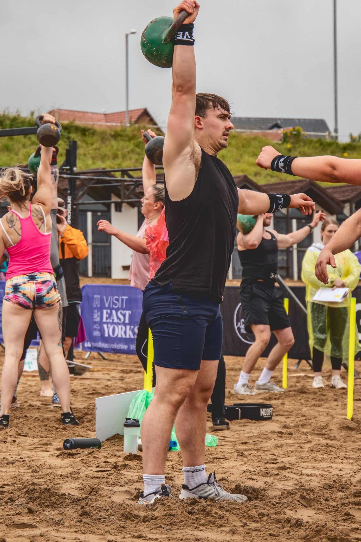 Athletes throw heavy kettlebells during a competitive outdoor fitness competition on sandy terrain.