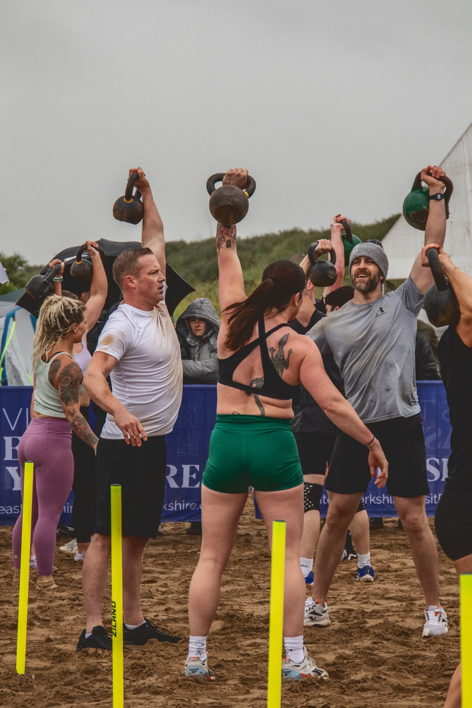 Athletes perform a synchronized kettlebell overhead press during an outdoor fitness competition on dirt ground.