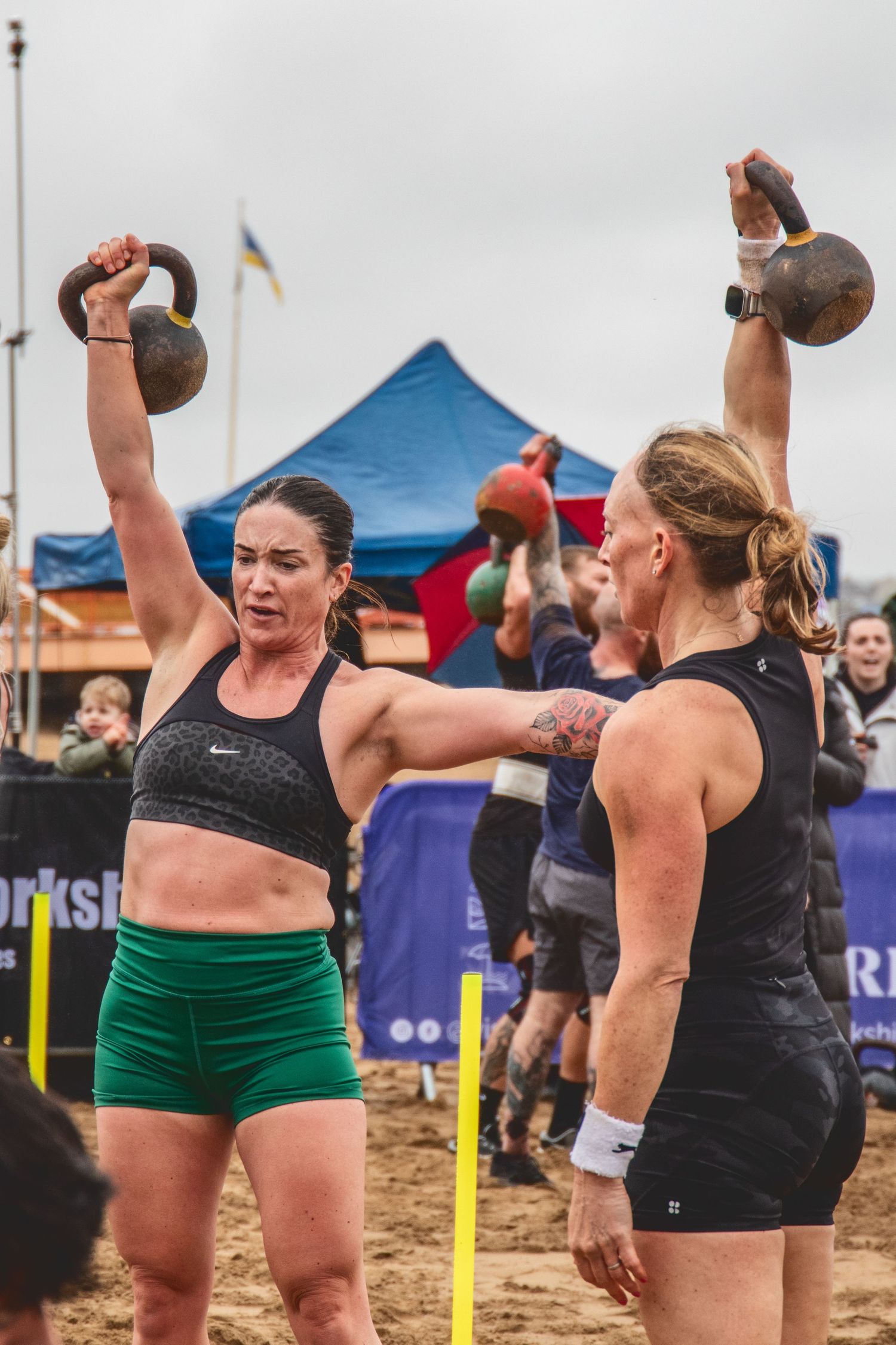 Athletes exercise with kettlebells during an outdoor competition on a sandy beach with tents in the background.