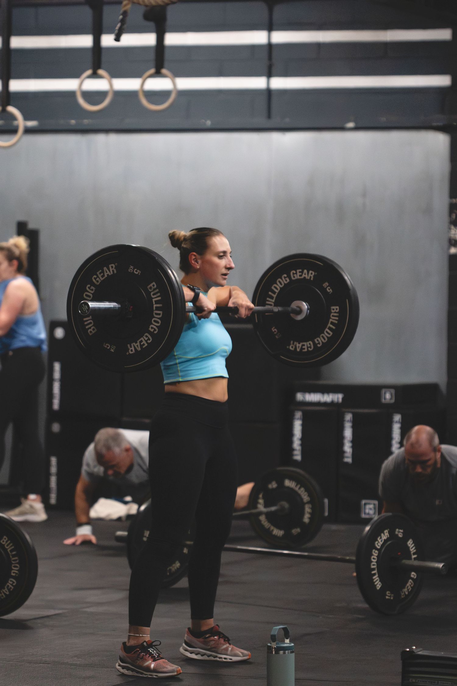 An athlete performs a barbell clean exercise in a CrossFit gym with gymnastic rings visible overhead.