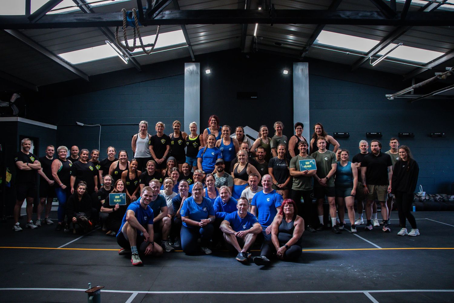 Large group photo of people wearing black and blue t-shirts gathered together in an indoor athletic facility.