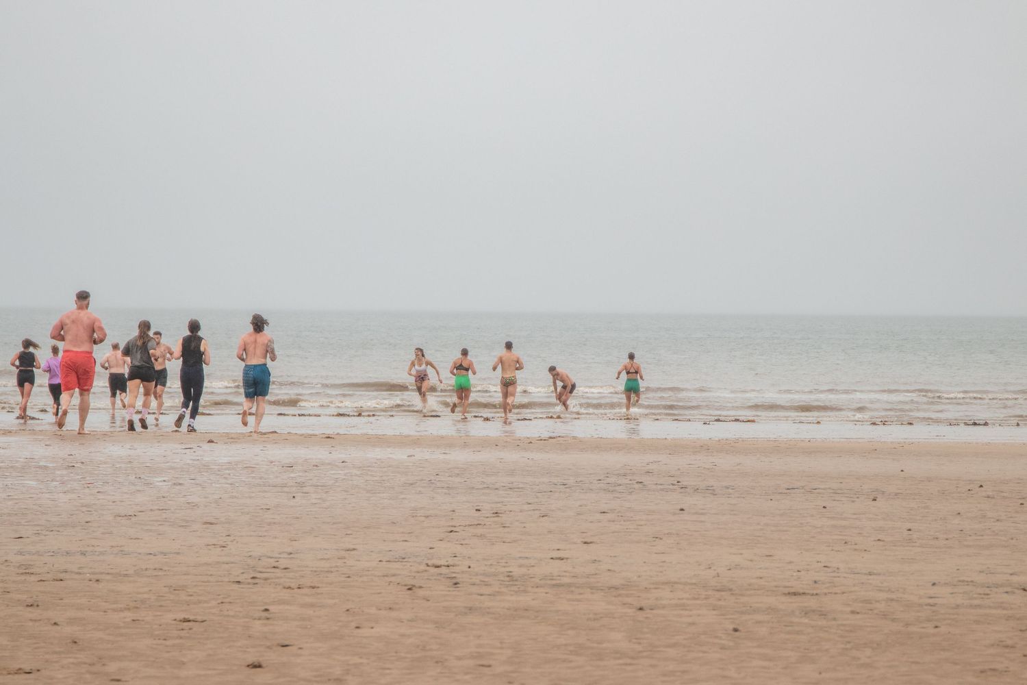 A group of people jogging along a misty beach shoreline on an overcast day.