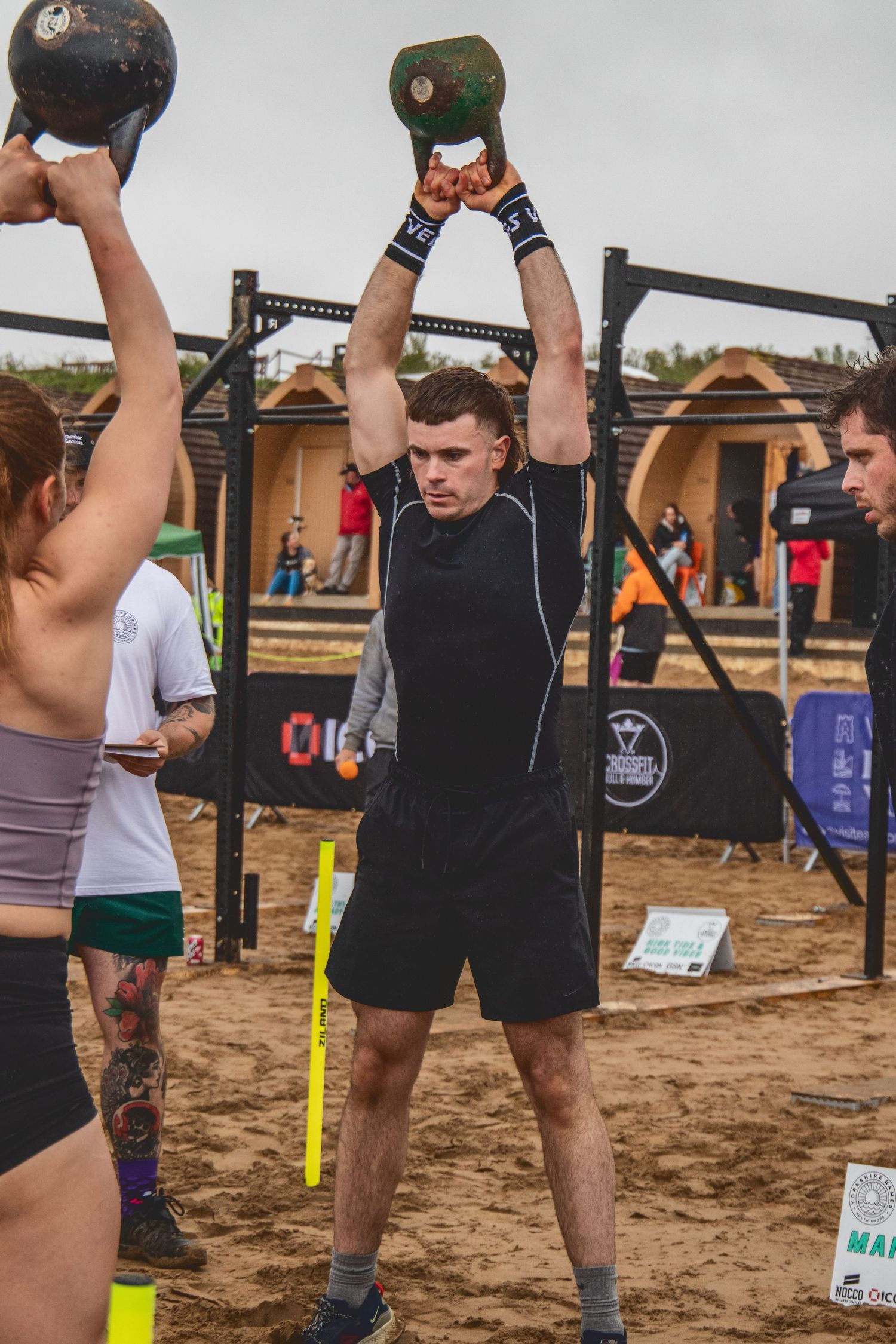 Athletes perform overhead weightlifting exercises at an outdoor competition on sandy terrain.