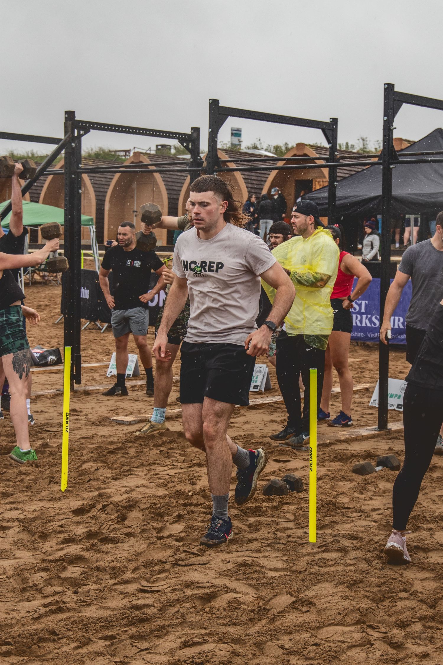 Athletes compete in an outdoor fitness competition with pull-up bars and equipment on sandy ground at a CrossFit event.