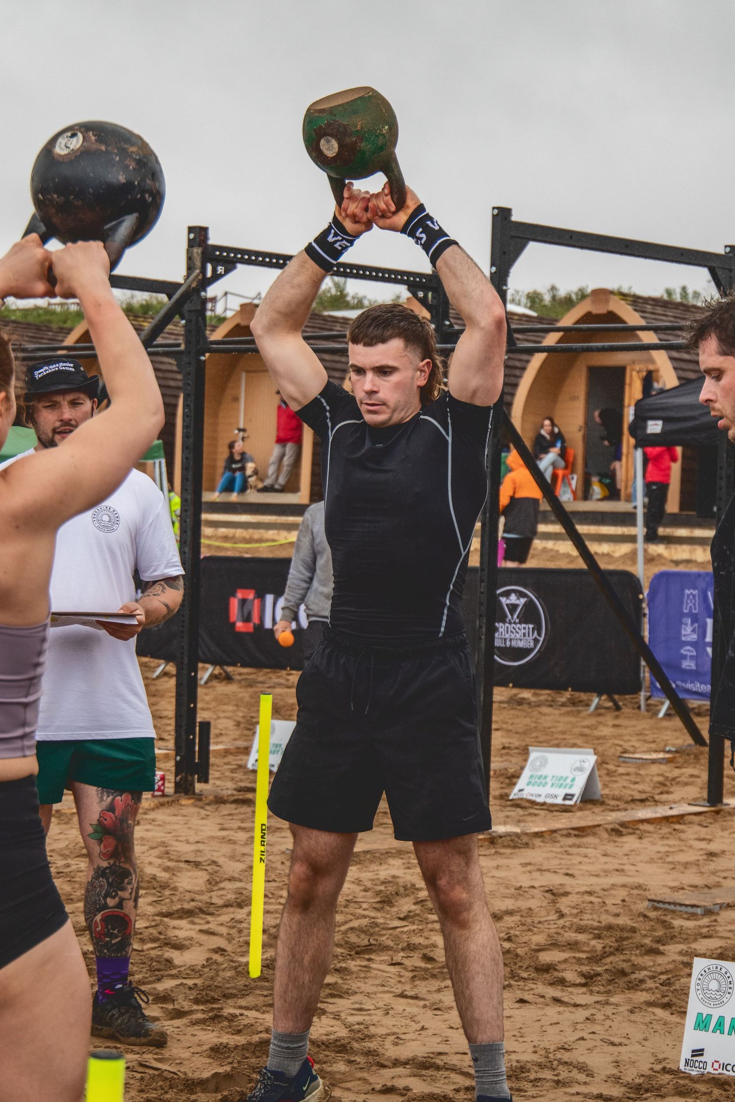 Athletes perform kettlebell overhead presses during an outdoor CrossFit competition on sandy terrain.