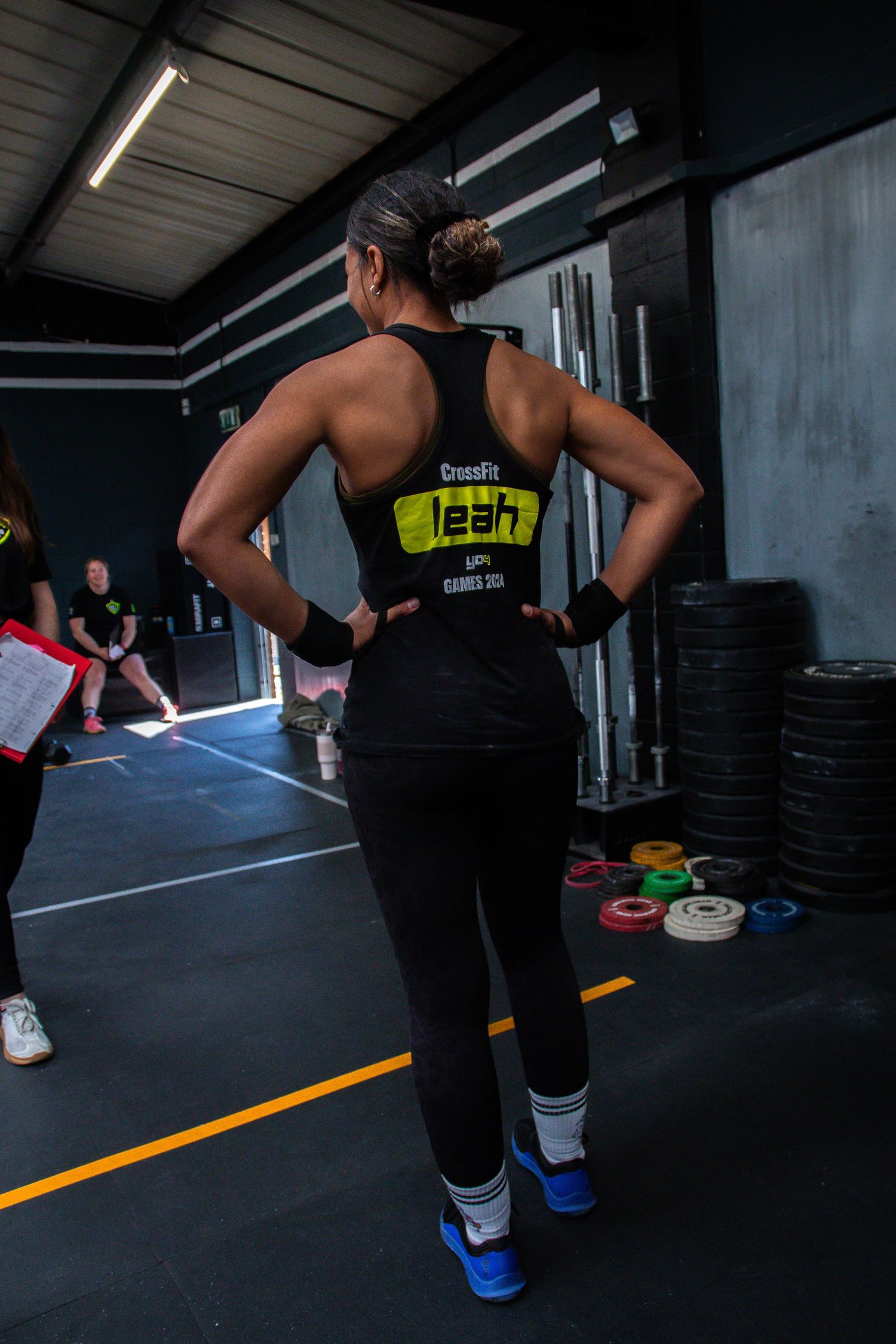 Athletic person in black workout attire stands in CrossFit gym with weight plates and training equipment visible.