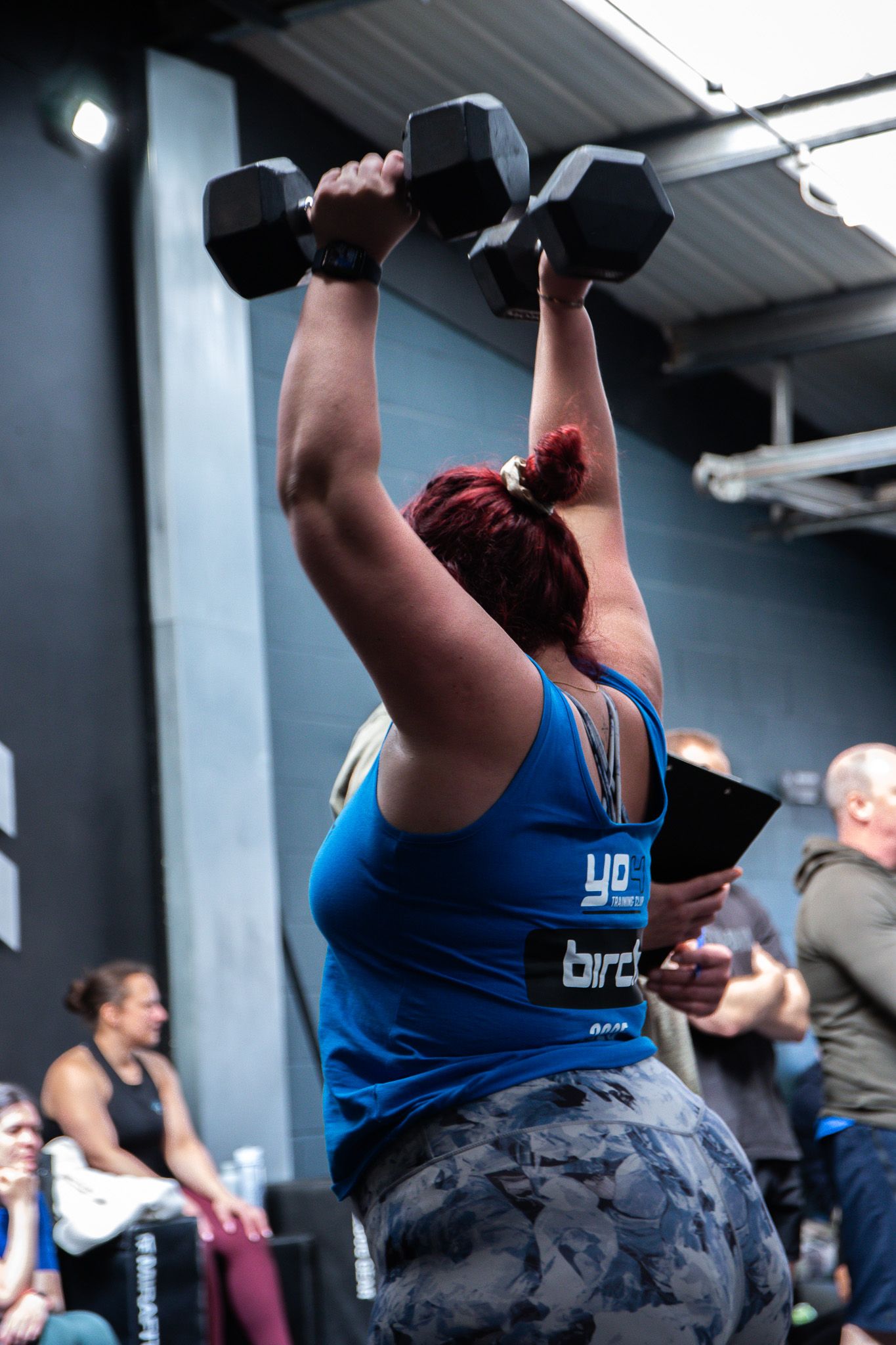 Athlete in blue tank top performs overhead dumbbell press during workout in gym with camouflage leggings.