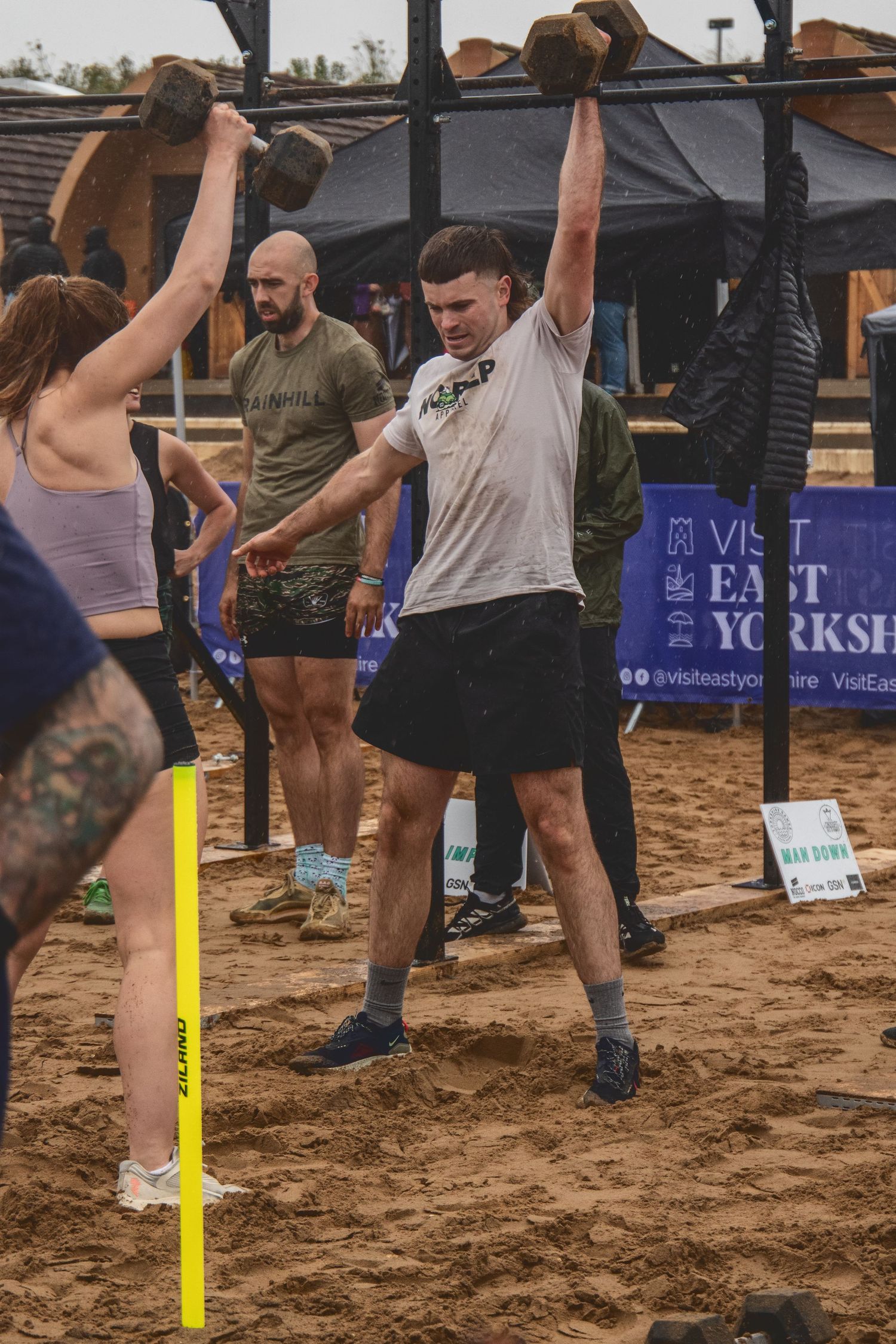 Athletes lift heavy weights during an outdoor CrossFit competition on sandy terrain with event banners visible in background.