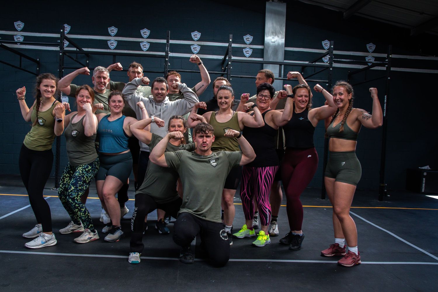 A group poses together in athletic wear at a fitness facility showing enthusiasm and flexing their muscles.