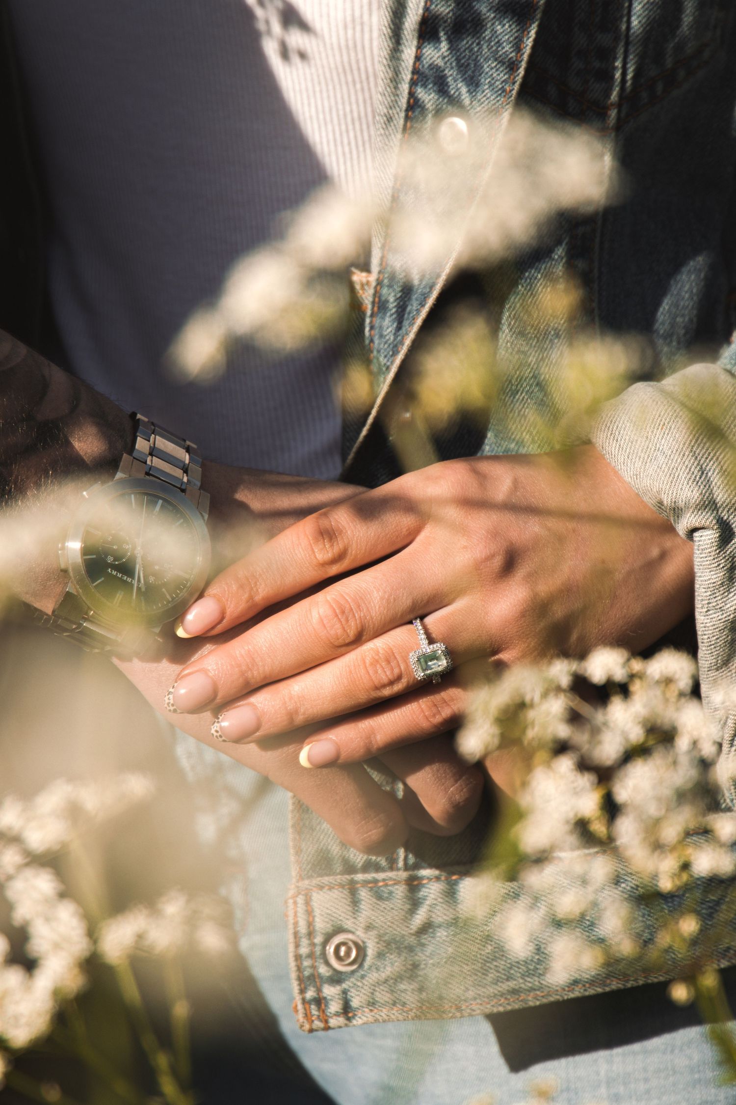 Close-up of engagement rings on hands with soft focus flowers in the background.