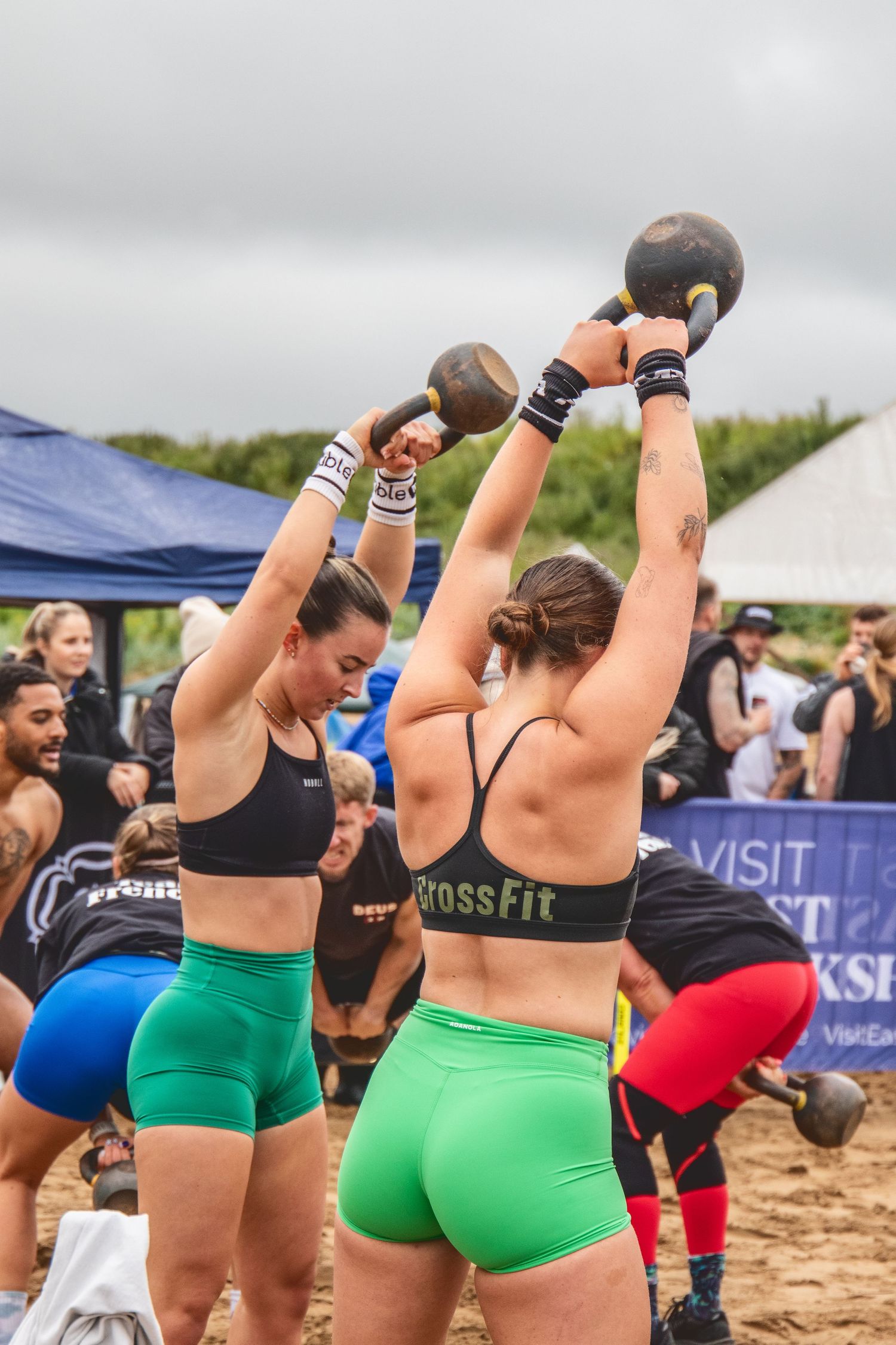 Athletes compete in a CrossFit competition on the beach lifting kettlebells while wearing bright colored workout gear.