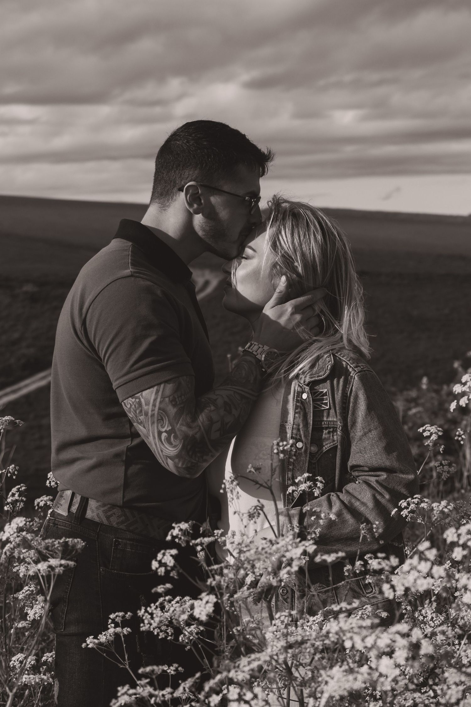 A romantic black and white photo of a couple embracing in a flower field under cloudy skies.