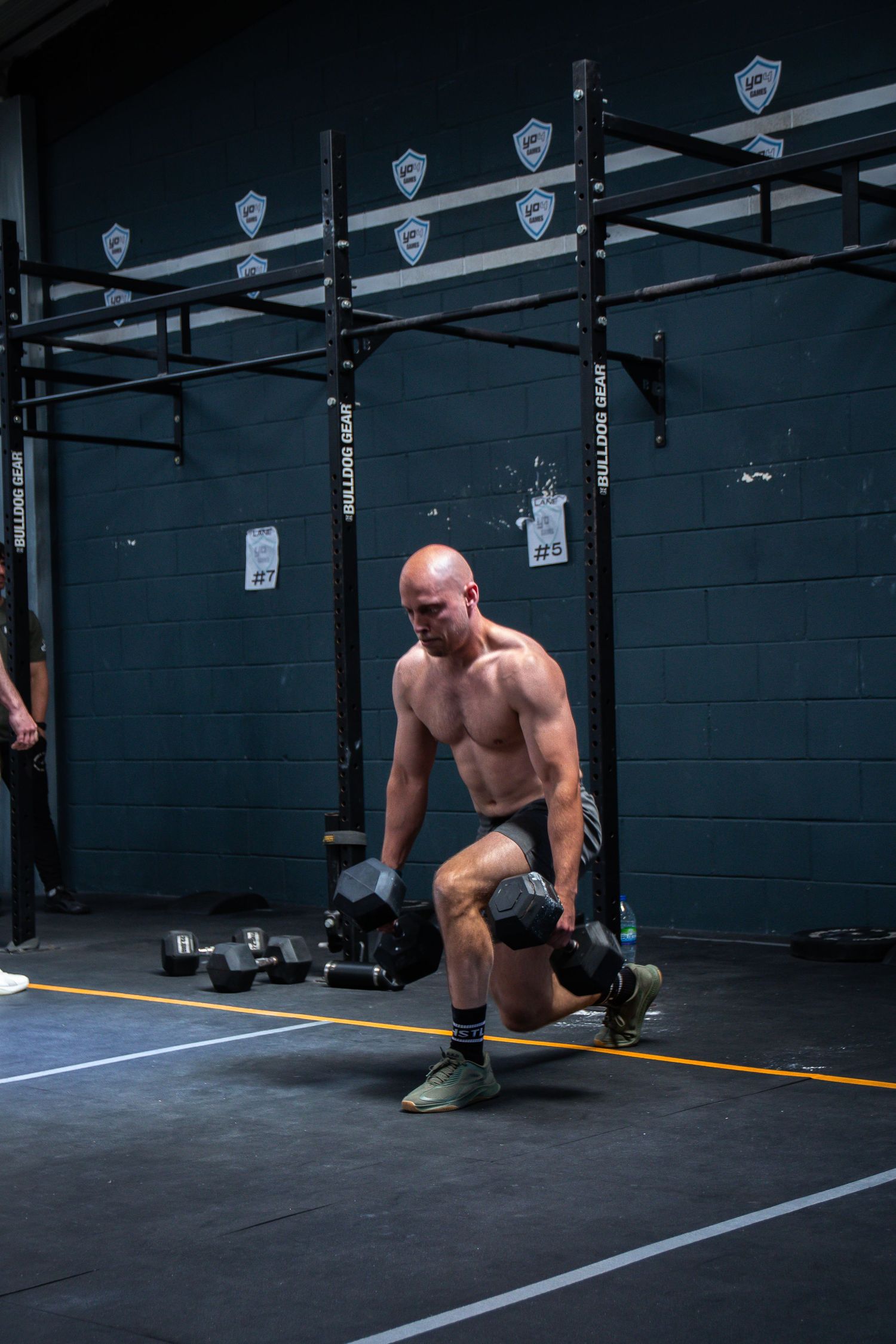 A shirtless athlete performs dumbbell lunges at a CrossFit gym with pull-up bars and dark walls visible in background.