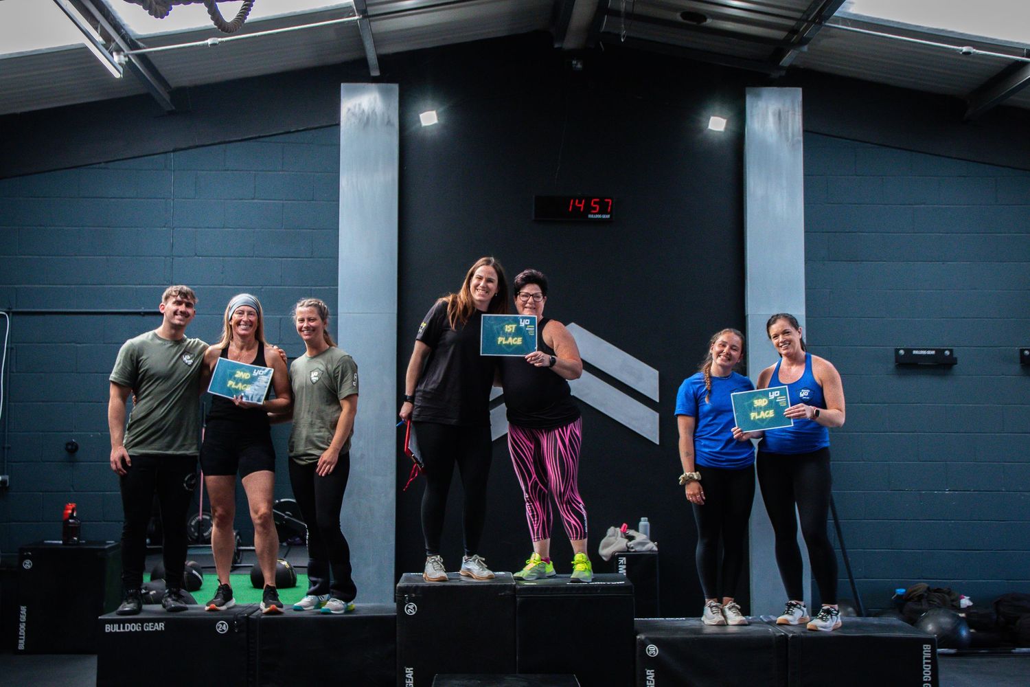 A group of fitness enthusiasts pose together on a platform in a gym with dark blue walls.
