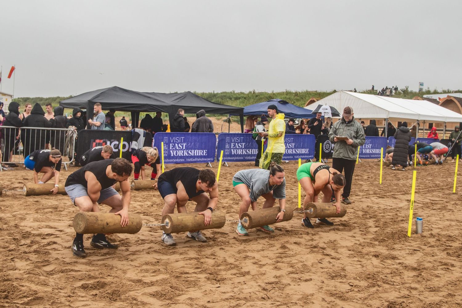Athletes compete in a beach log lifting competition during an outdoor sporting event with banners and spectators.