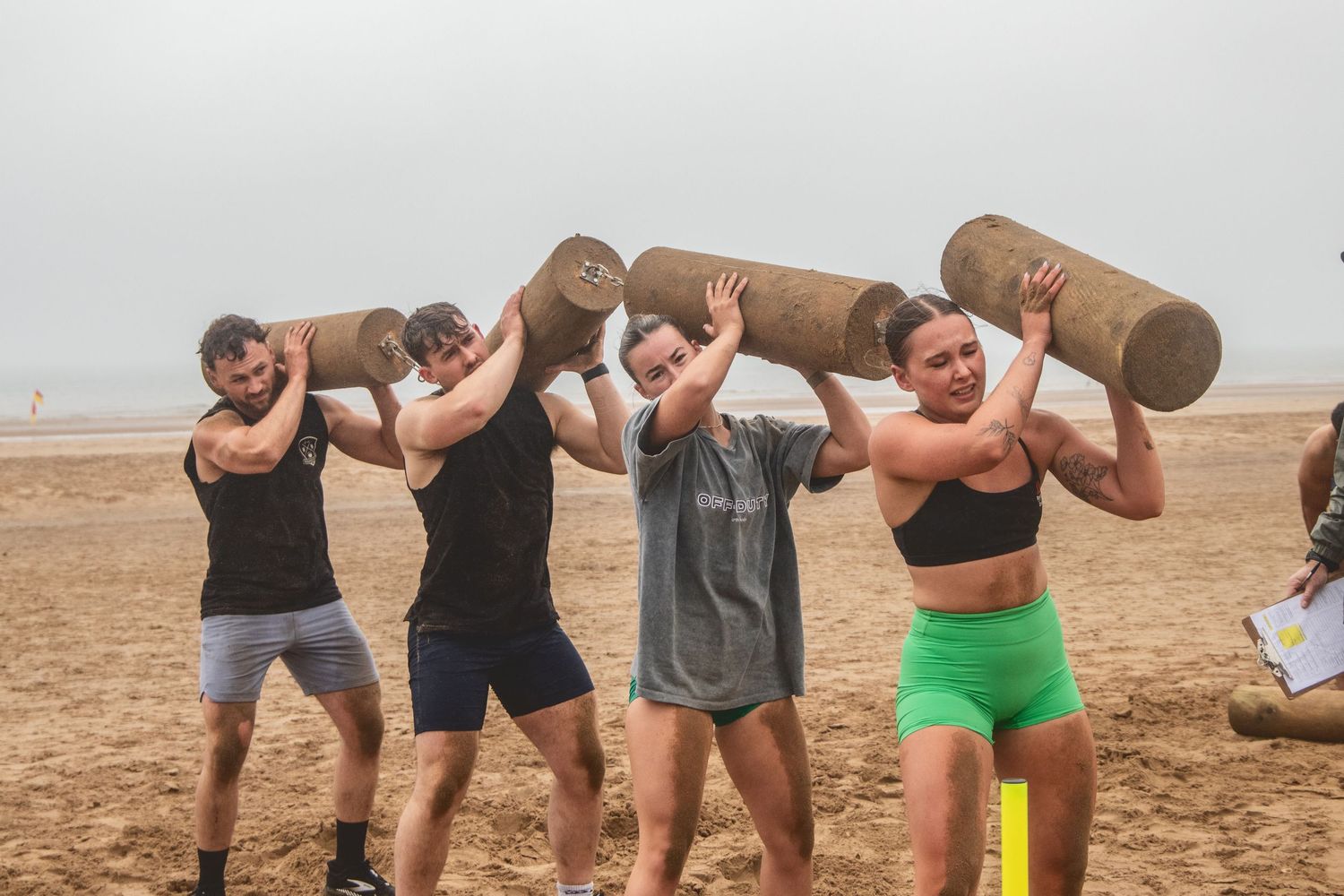 A group of fitness enthusiasts perform shoulder exercises with water jugs on a foggy beach during an outdoor workout.