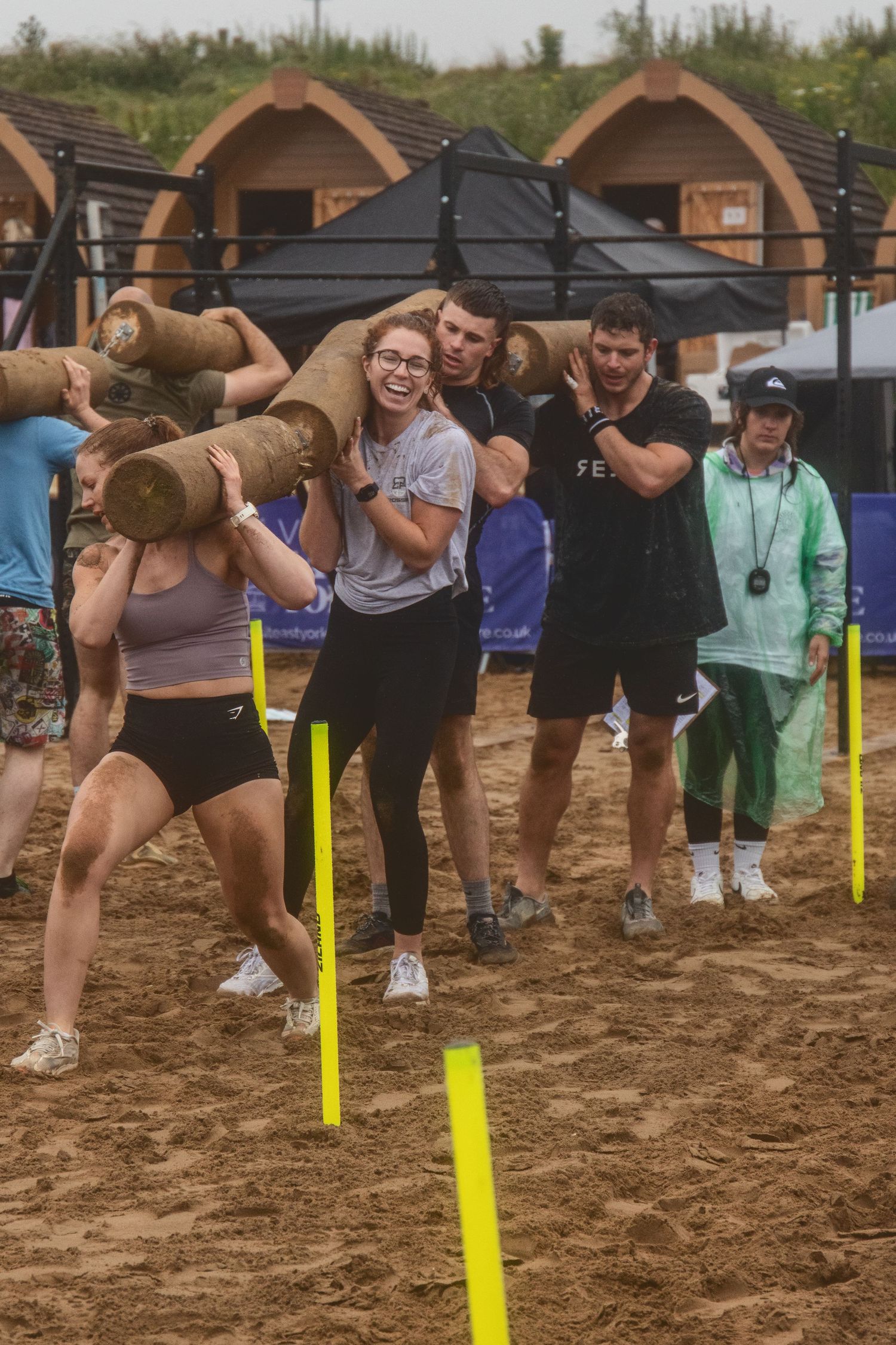 Group participates in a beach obstacle course race carrying large teddy bears past yellow markers in the sand.
