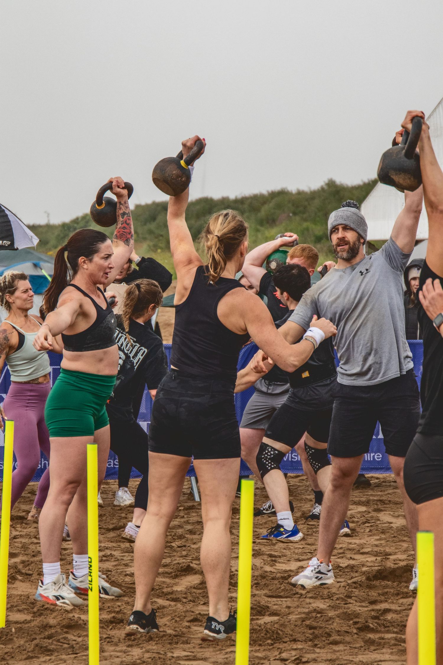 Athletes perform kettlebell exercises during an outdoor fitness competition on sandy terrain with yellow markers.