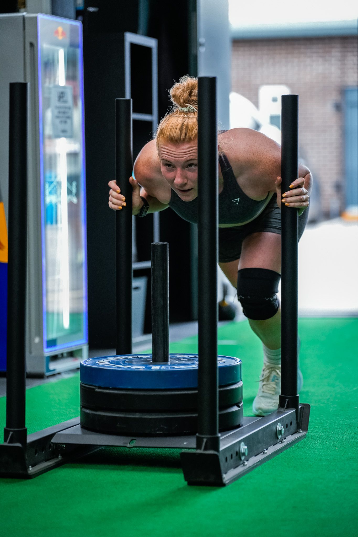 Person doing a burpee exercise on green turf flooring in a fitness facility with black equipment stands visible.