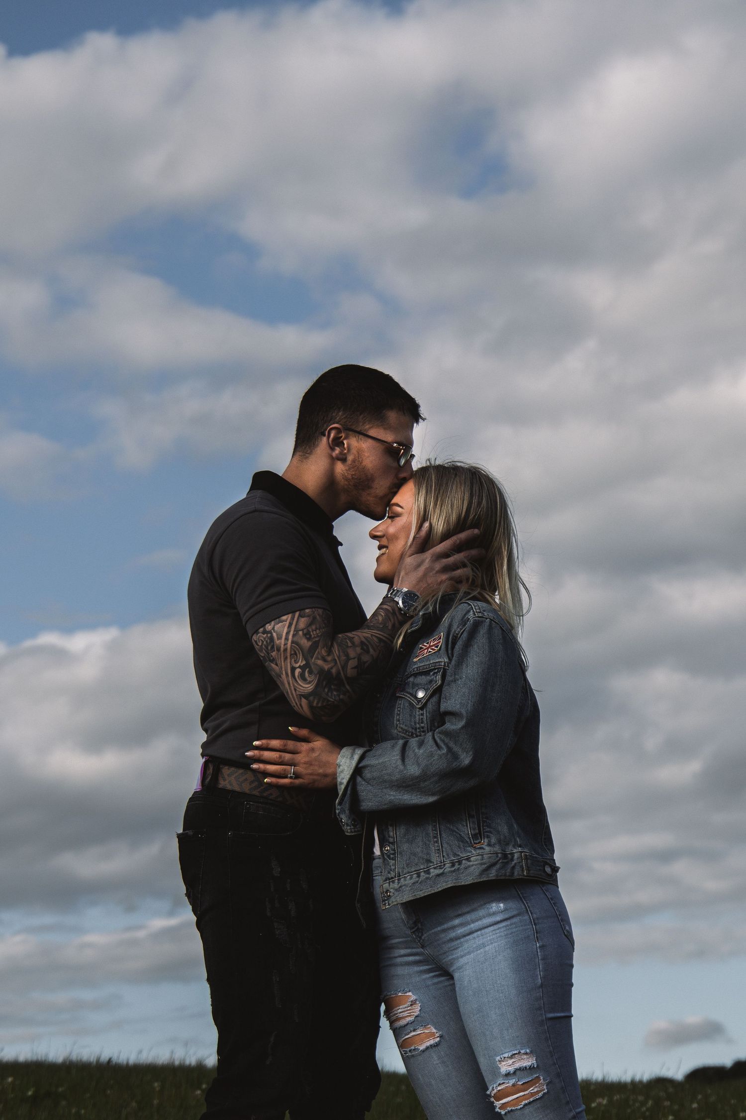 A romantic couple embraces in a field under cloudy skies wearing casual denim and black attire.
