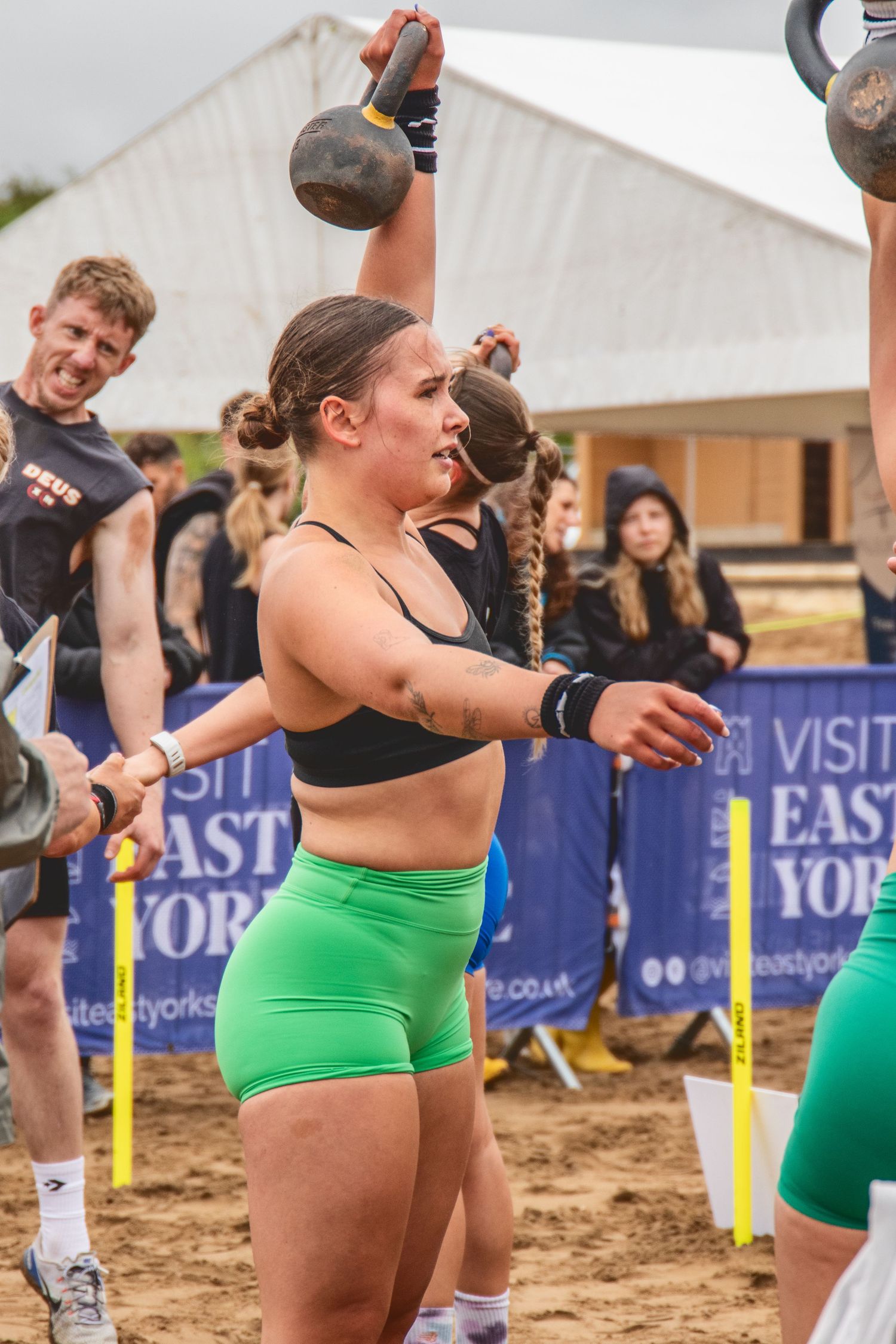 Athletes performing kettlebell exercises during a beach fitness competition on sandy terrain with event banners visible.