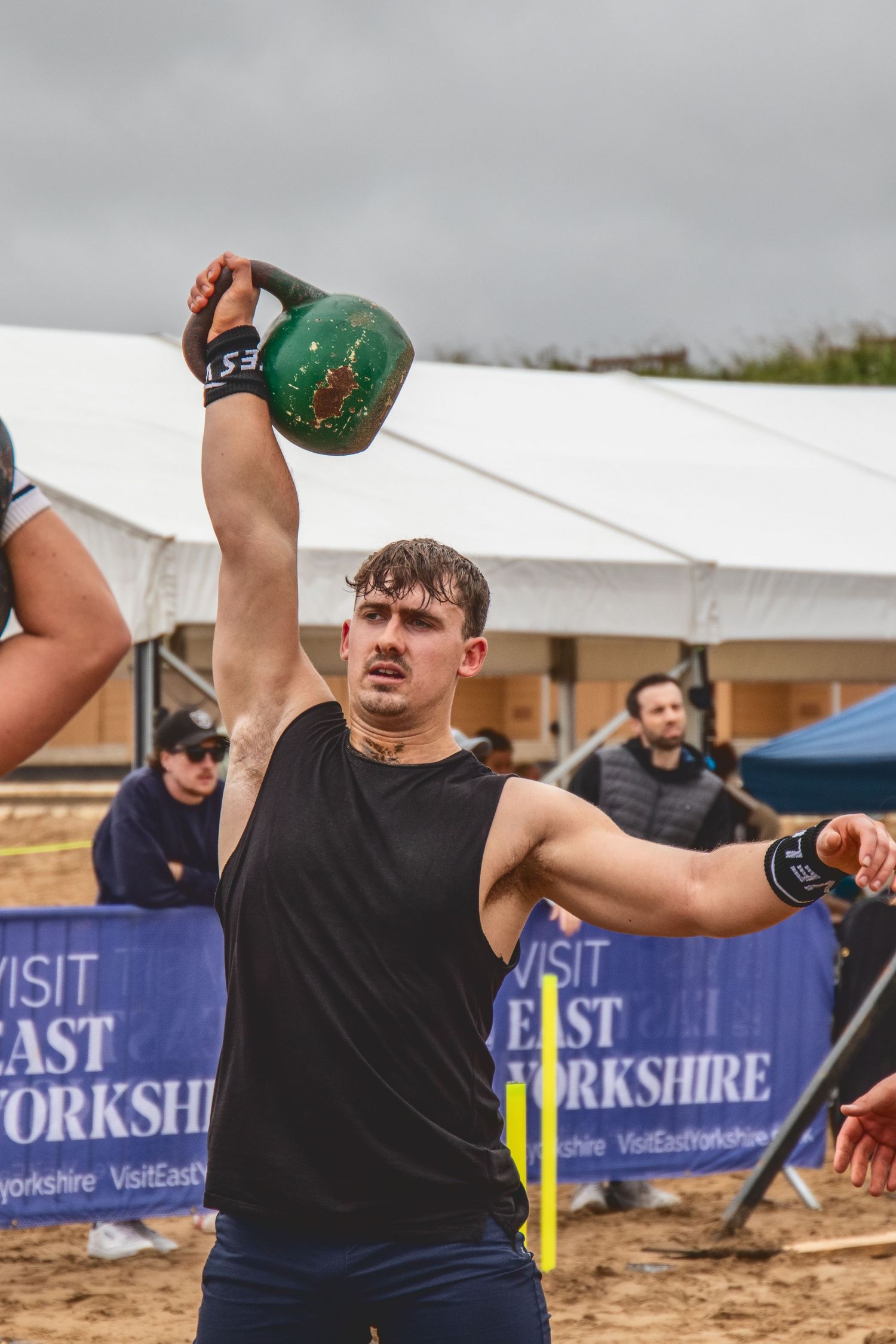 Three sequential images show an athlete practicing the kettlebell swing exercise at an outdoor competition venue.