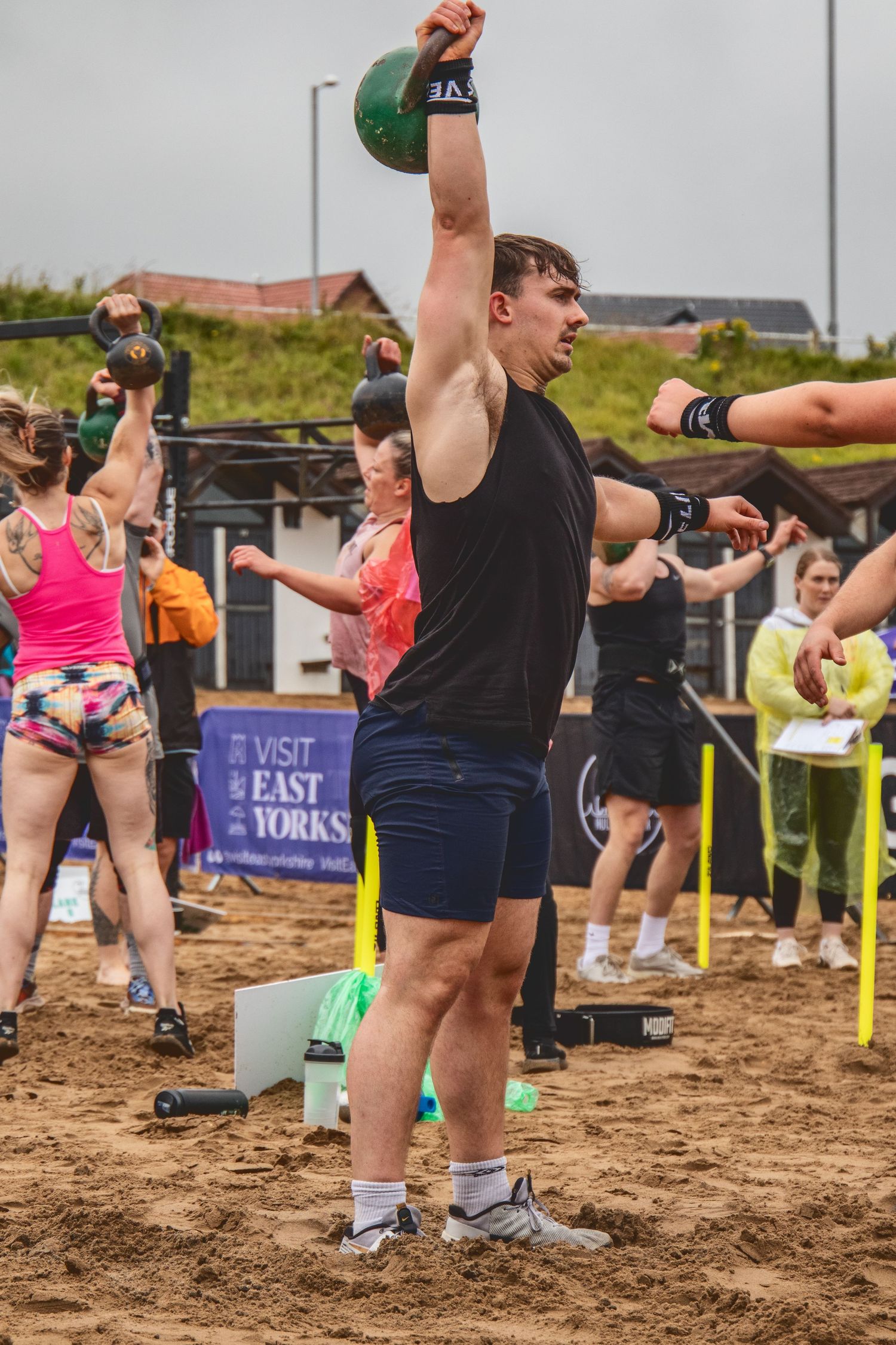 Athletes perform kettlebell exercises during an outdoor CrossFit competition on sandy ground.