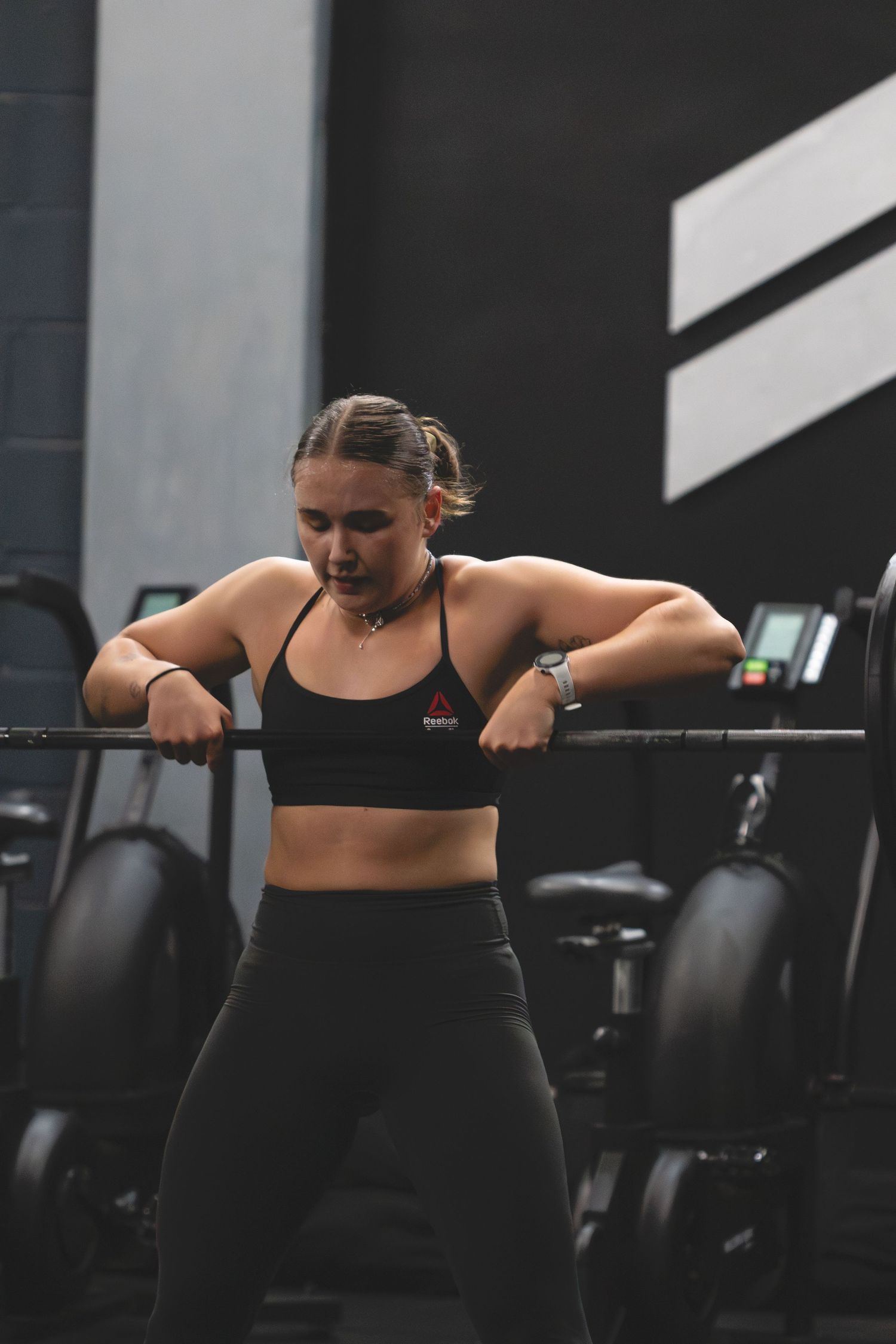 Person resting on barbell at gym wearing black sports bra and leggings during intense workout session.