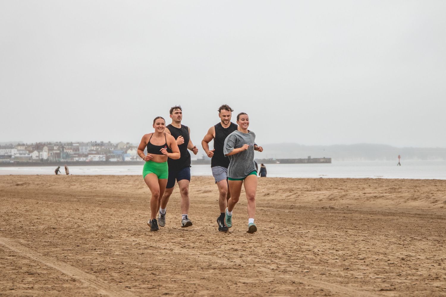 A group of athletes running together on a sandy beach with ocean and pier in the background.